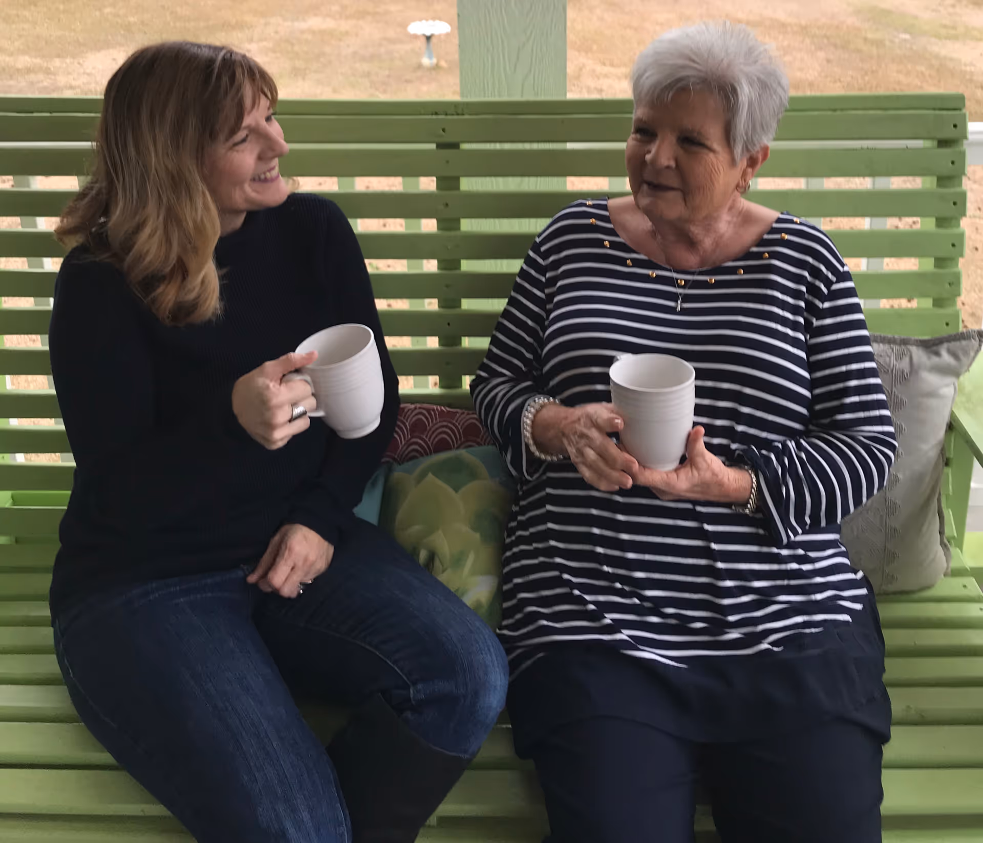 Two women sit on a green outdoor bench, chatting and holding white cups.