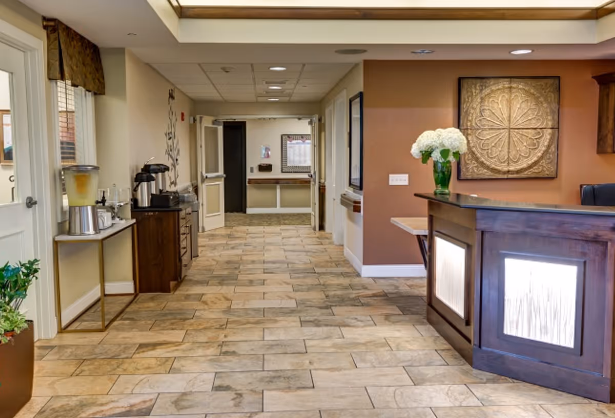 A spacious lobby with a wooden reception desk, tiled floor, beverage station, and flowers.