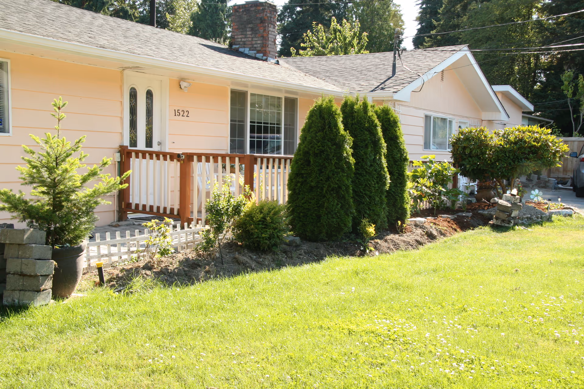 A single-story pastel house with a wooden front porch, lawn, and trimmed shrubs.