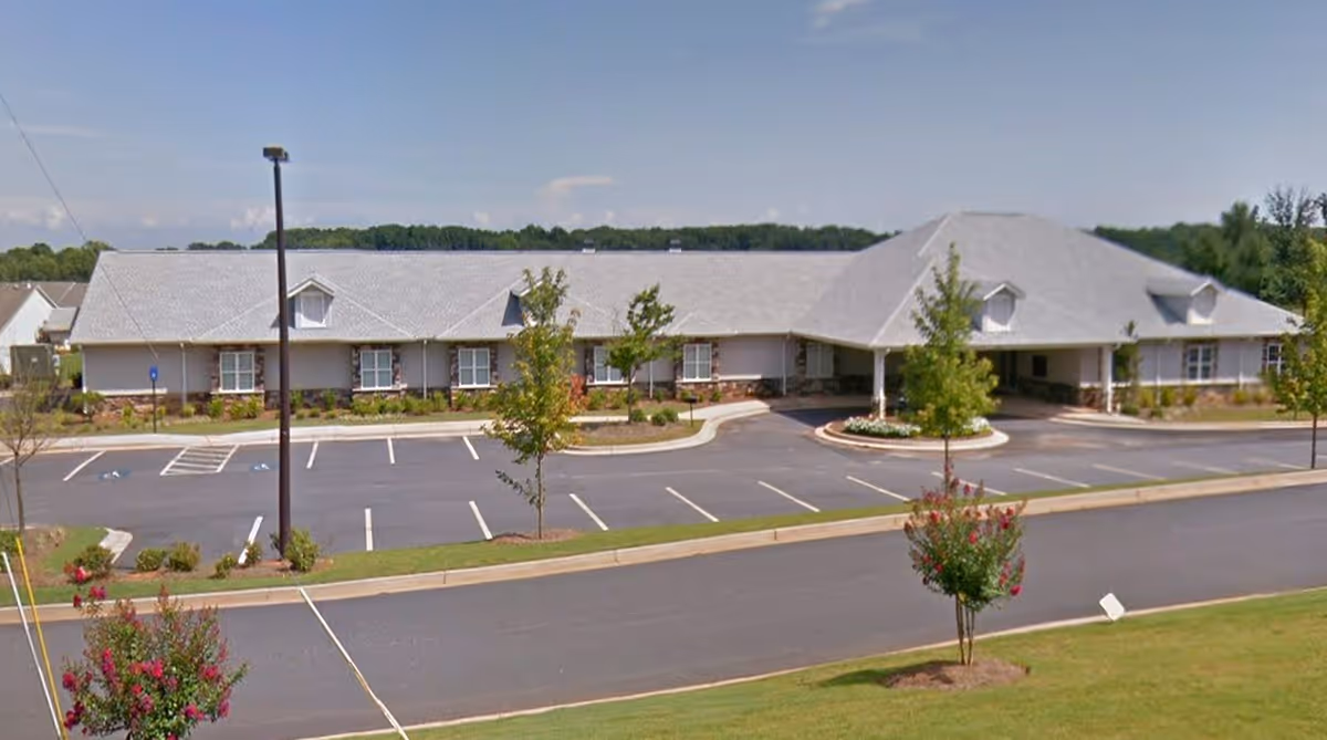 Single-story memory care facility building with a covered main entrance, parking lot, and landscaped grounds under a clear sky.