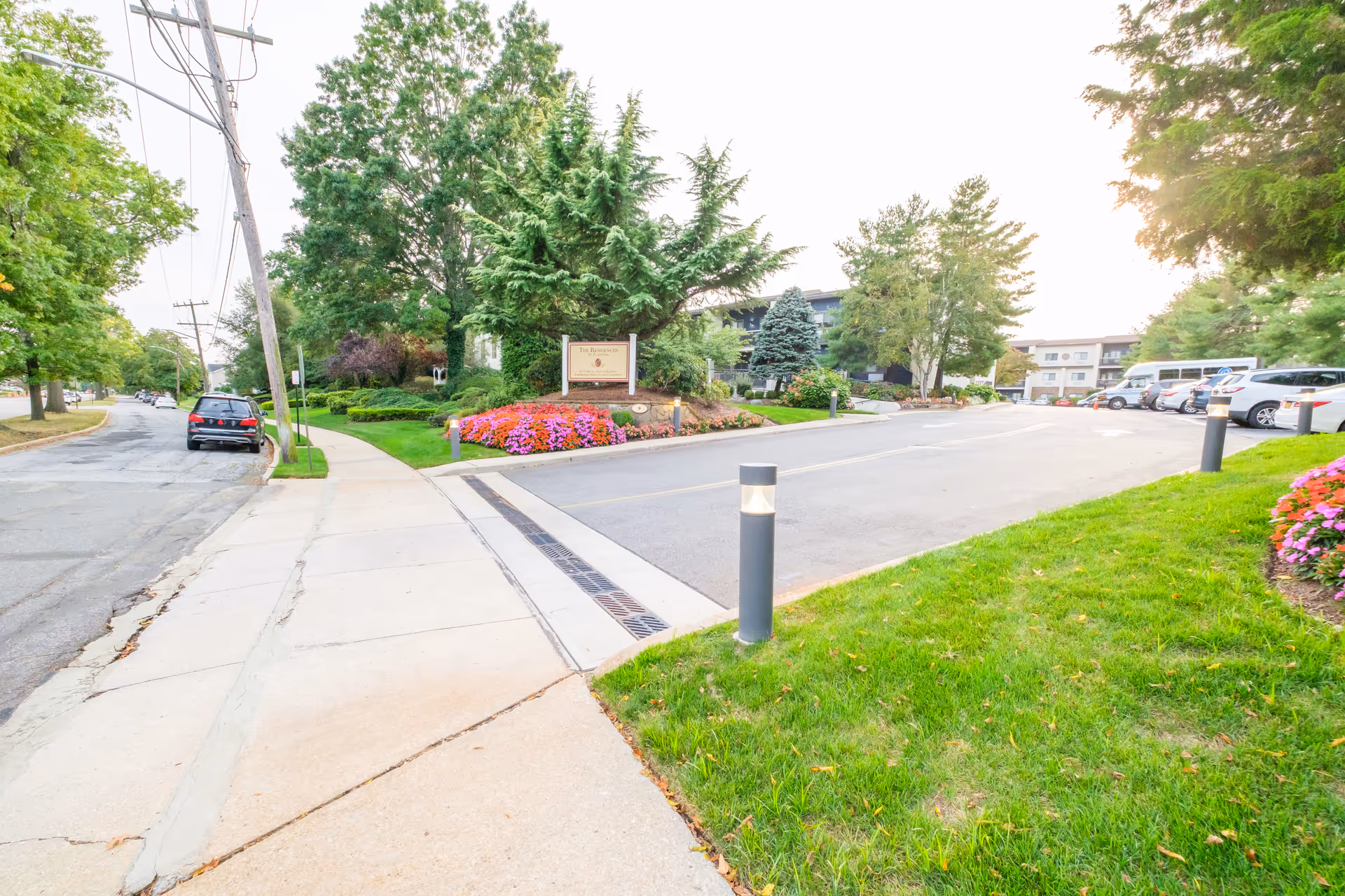 Landscaped front driveway and entrance of a senior living residence with a sign, flower beds, and parked cars.