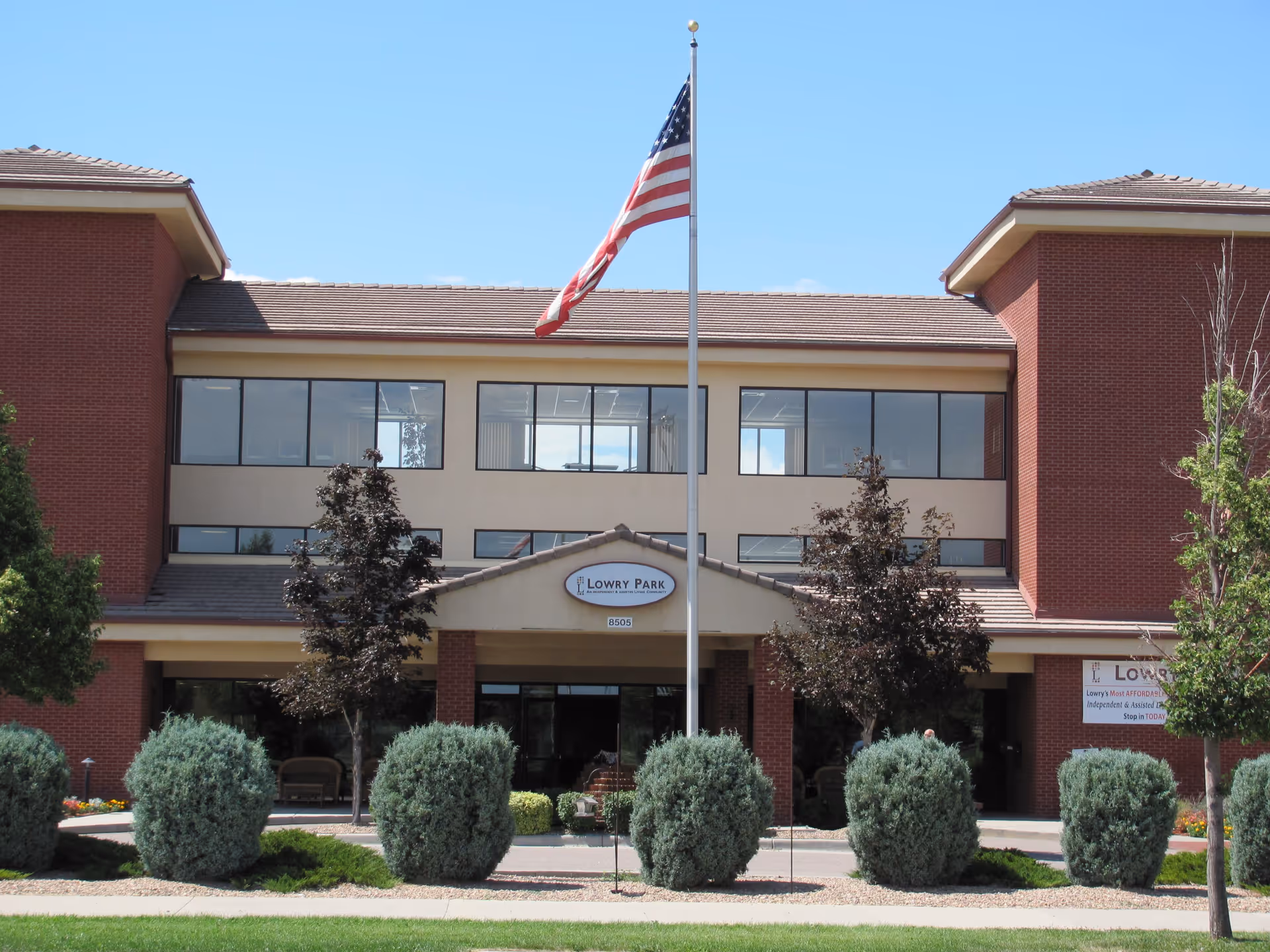 Front exterior view of the Lowry Park facility building with a flagpole displaying the American flag in the center. The building has two stories with large windows and red brick accents on the sides. There are neatly trimmed bushes and small trees in front of the entrance.