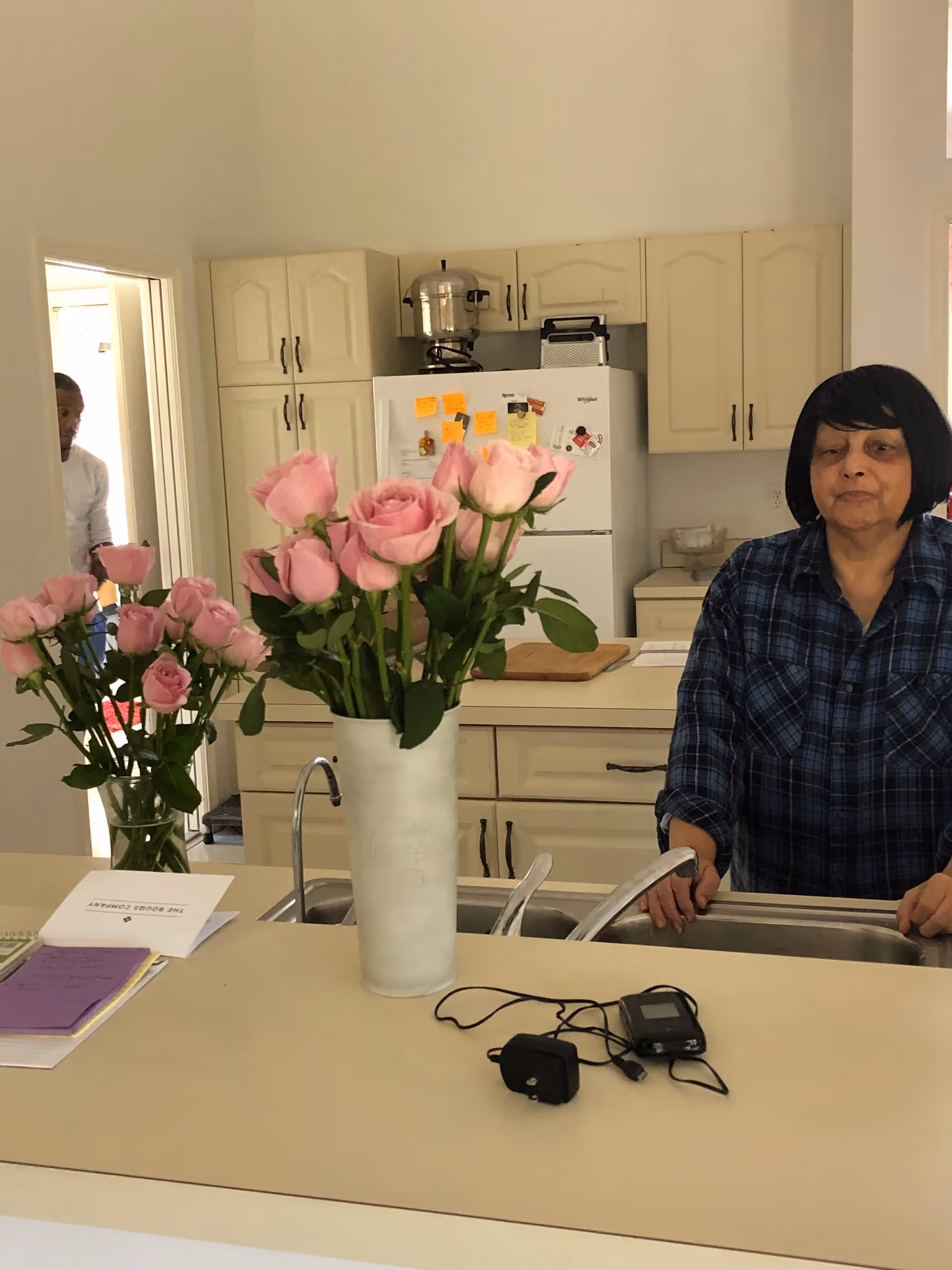 Woman stands at a kitchen counter with pink roses and a refrigerator in the background.