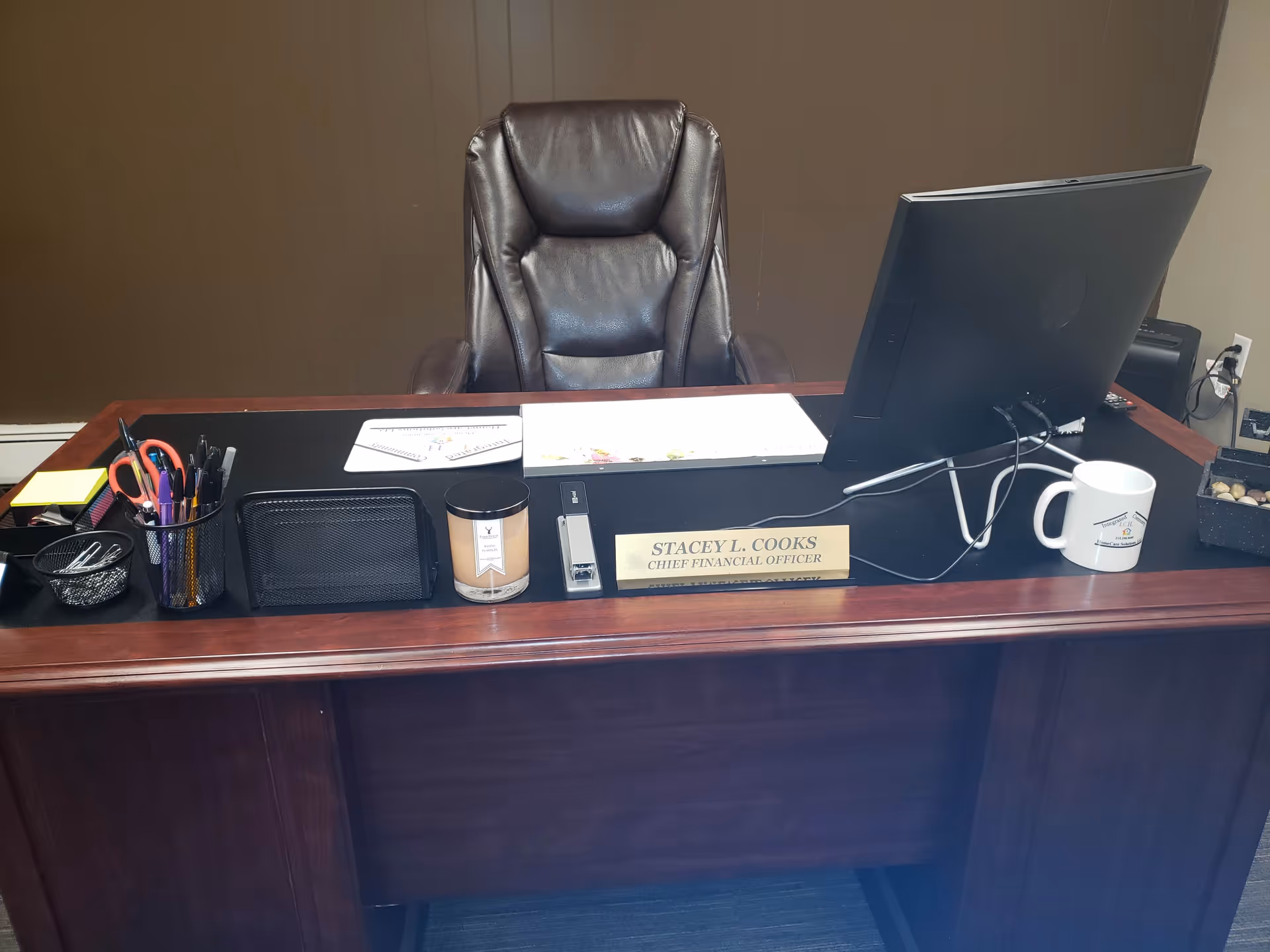 Executive desk setup in an office, featuring chair, computer, nameplate, mug, and supplies.