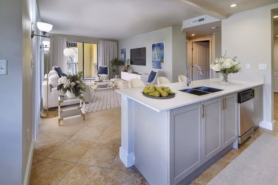 Modern open-concept living space with a kitchen island featuring a sink and a bowl of pears. The kitchen island has gray cabinets and a white countertop with a vase of white flowers. The living room area includes a white sofa, two blue armchairs, a coffee table, a TV mounted on the wall, and decorative artwork. Large sliding glass doors with curtains lead to an outdoor balcony.