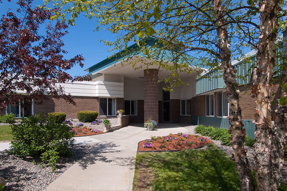 Entrance area of Medilodge of Sault Ste. Marie facility showing a covered walkway supported by a brick column, surrounded by landscaped garden beds with flowers, green grass, and trees under a clear blue sky.