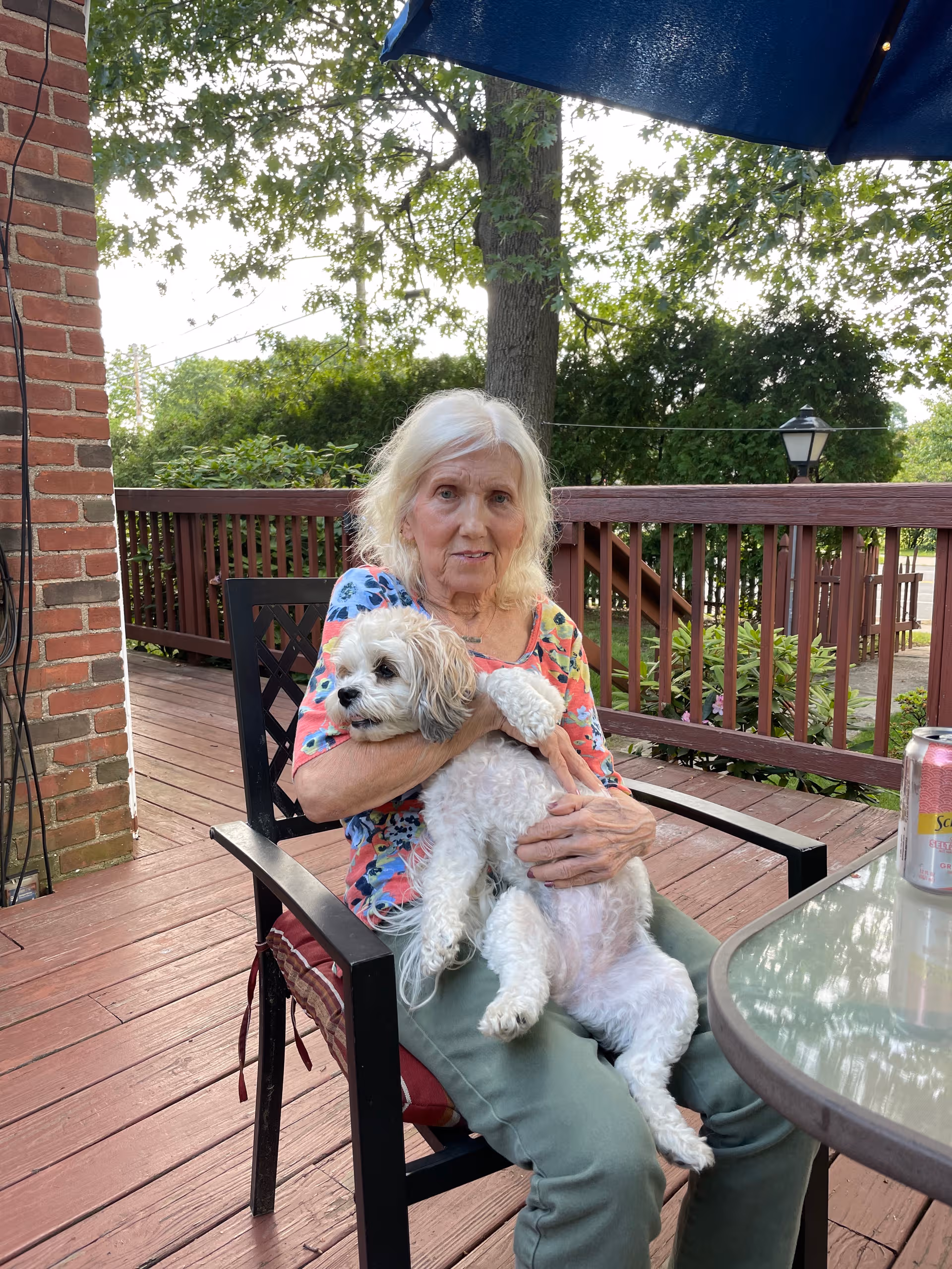 Elderly woman sits on a wooden outdoor deck, holding a white dog under a blue umbrella.