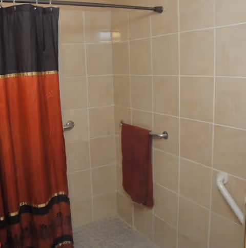 Tiled bathroom corner with a red and black shower curtain, a red towel on a wall-mounted towel bar, and a safety grab bar.