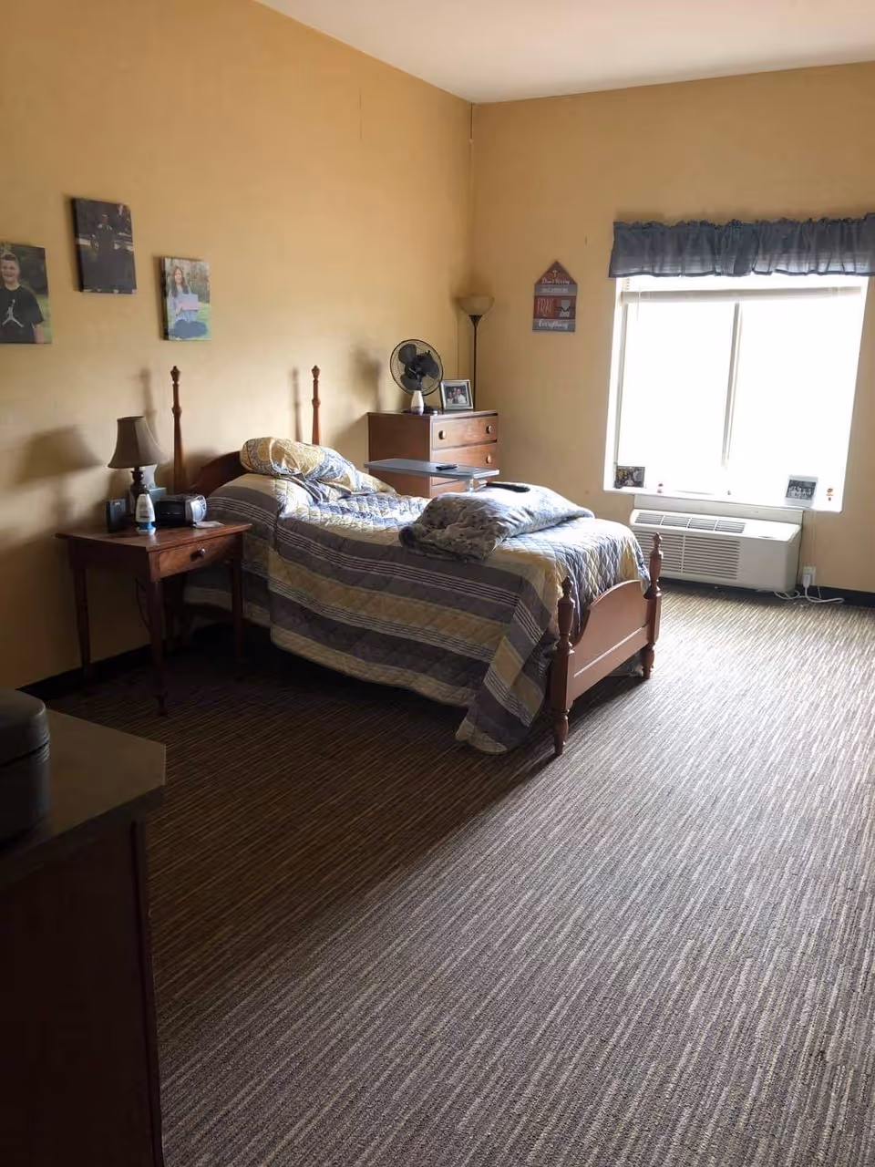 A single-occupancy bedroom with a twin bed, nightstand and lamp, dresser, window with an air conditioner, and photos on the walls.