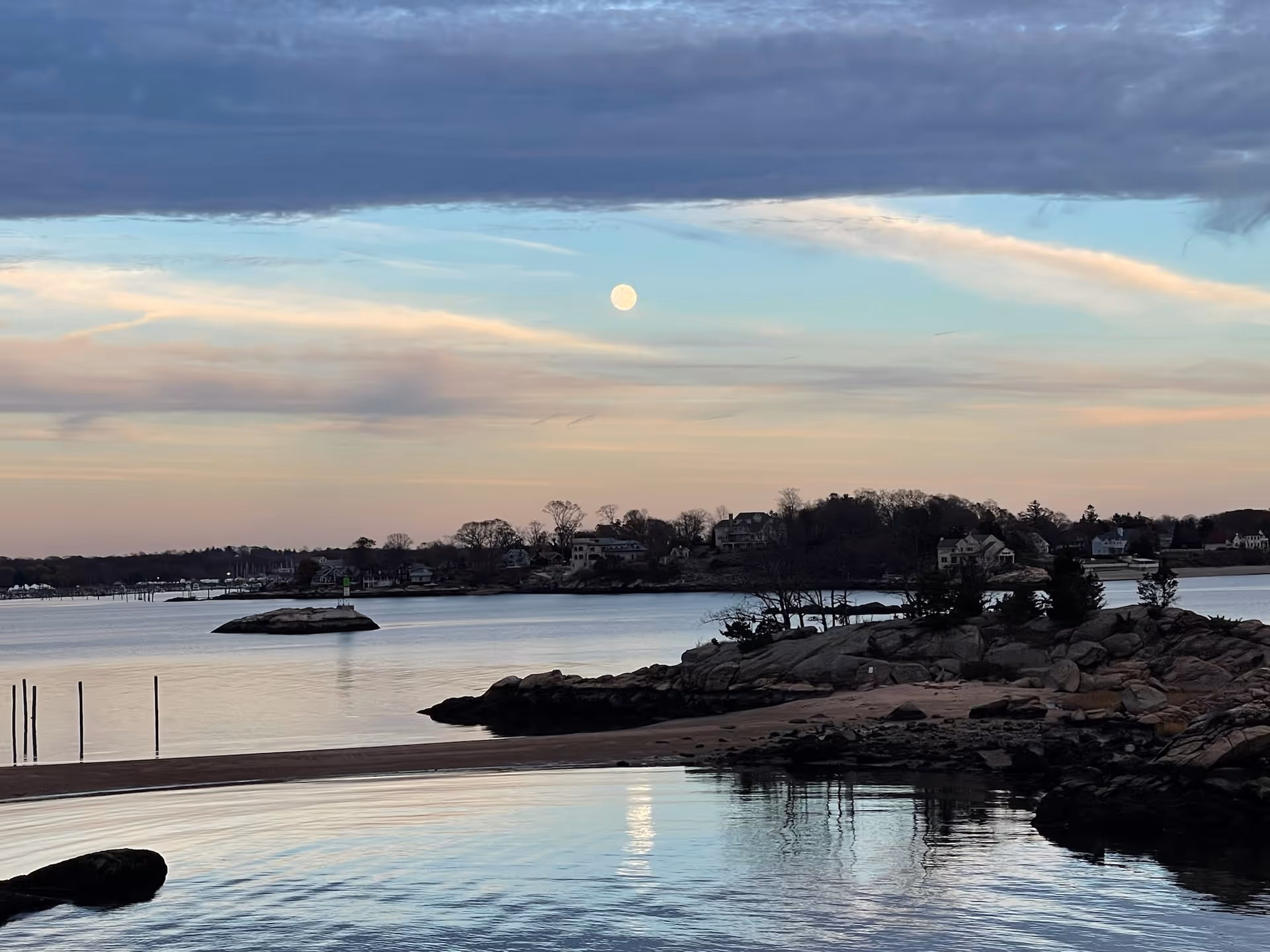 Moonlit coastal scene with rocky shoreline, calm water, and distant houses.
