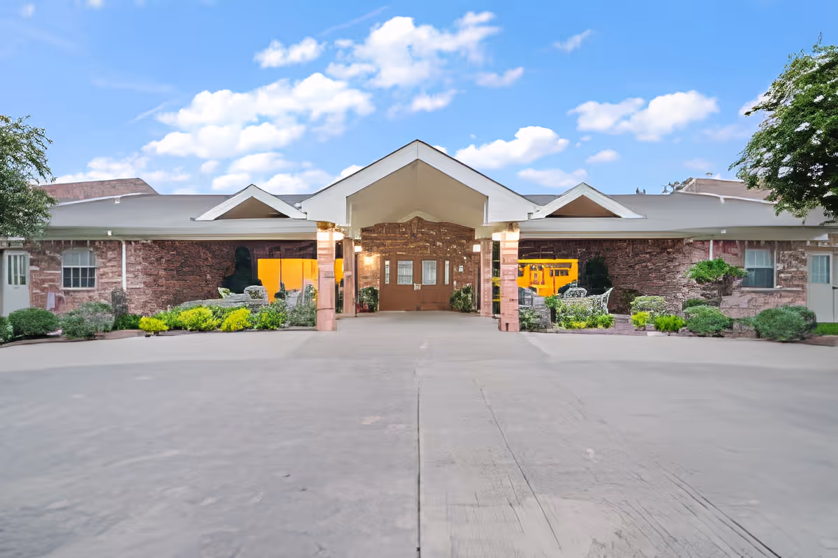 Front exterior view of Morada Victoria facility showing a single-story building with a covered entrance, brick walls, windows, and landscaped bushes under a blue sky with scattered clouds.