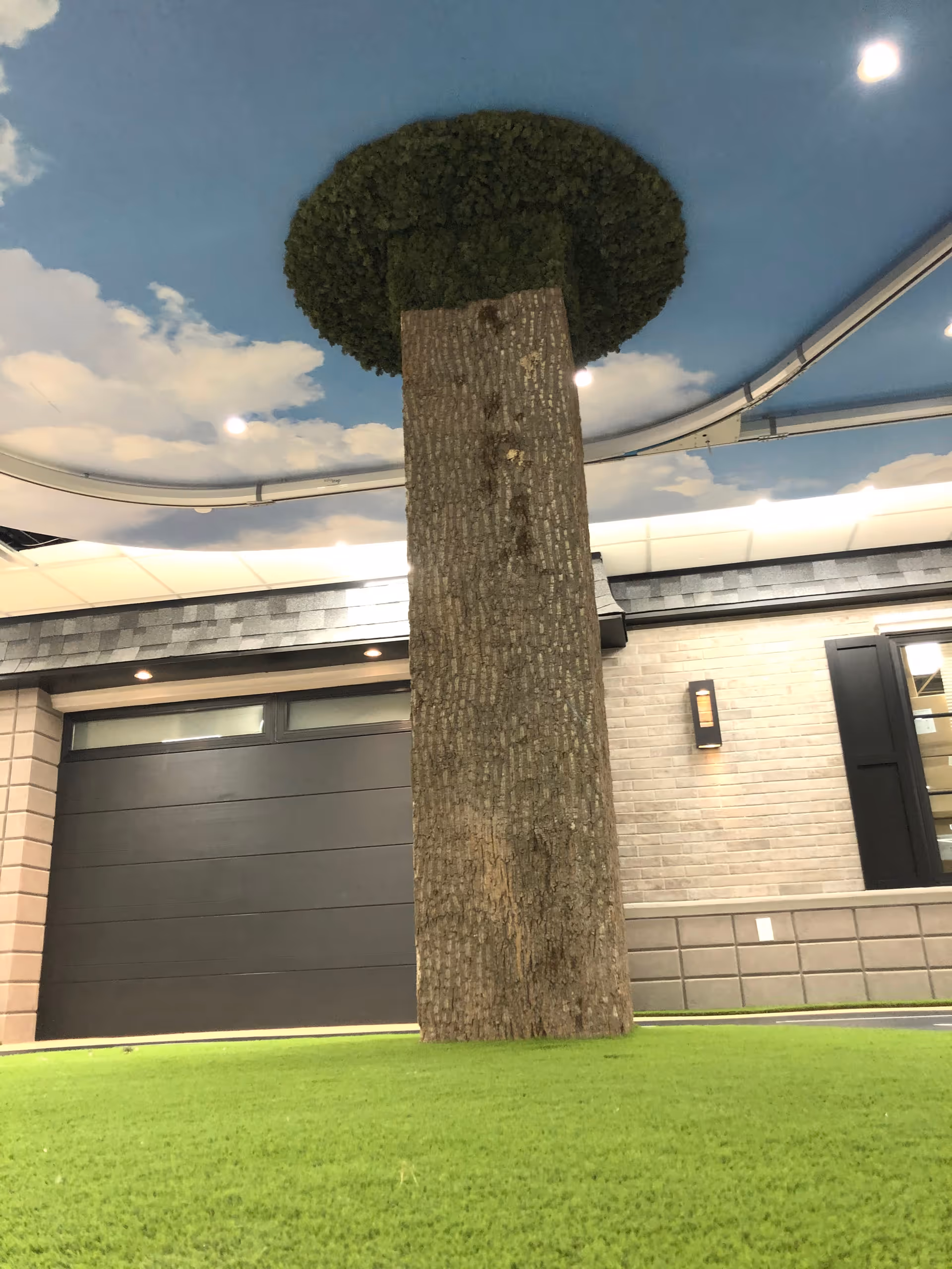 Indoor area with artificial green grass flooring, a large artificial tree trunk extending from the floor to the ceiling, and a ceiling painted with a blue sky and white clouds. The background includes a wall with a garage door and window shutters.