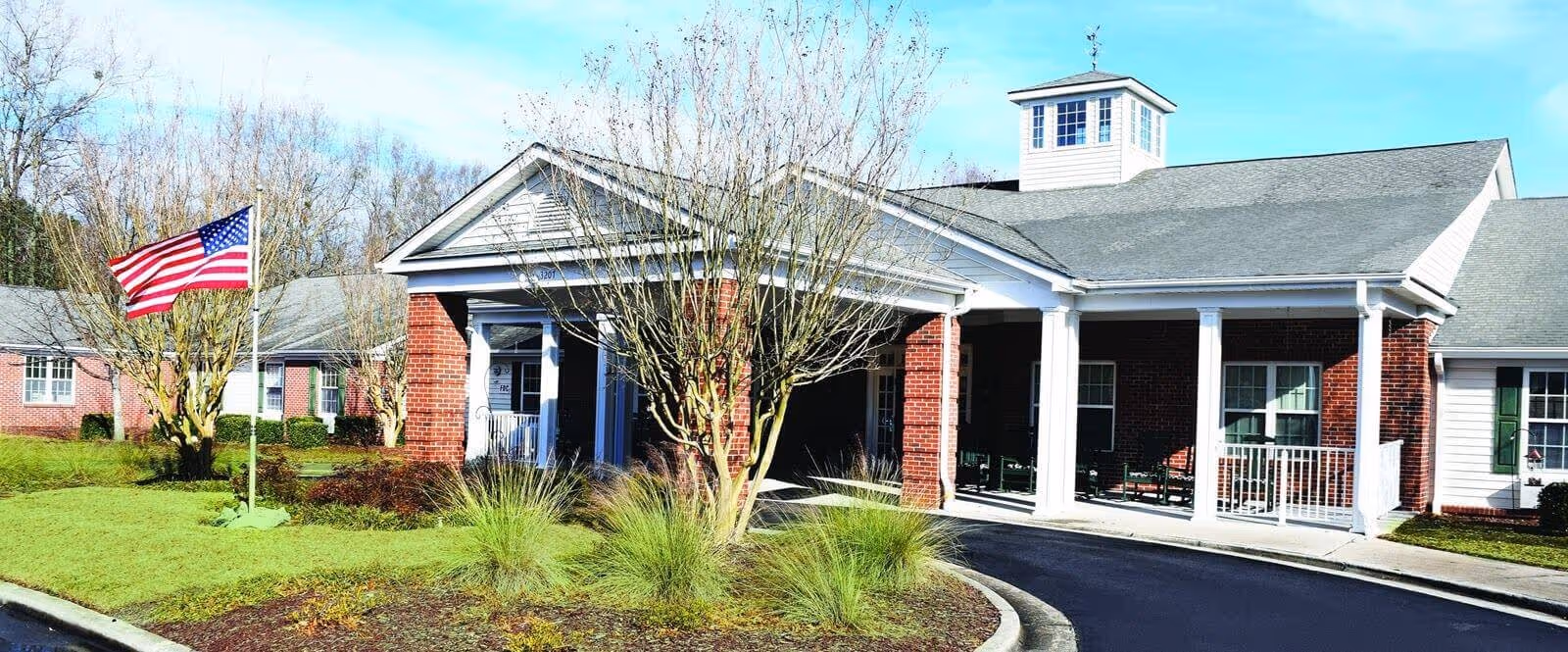Front entrance of a brick senior living building with a covered porch, American flag, and landscaped grounds.