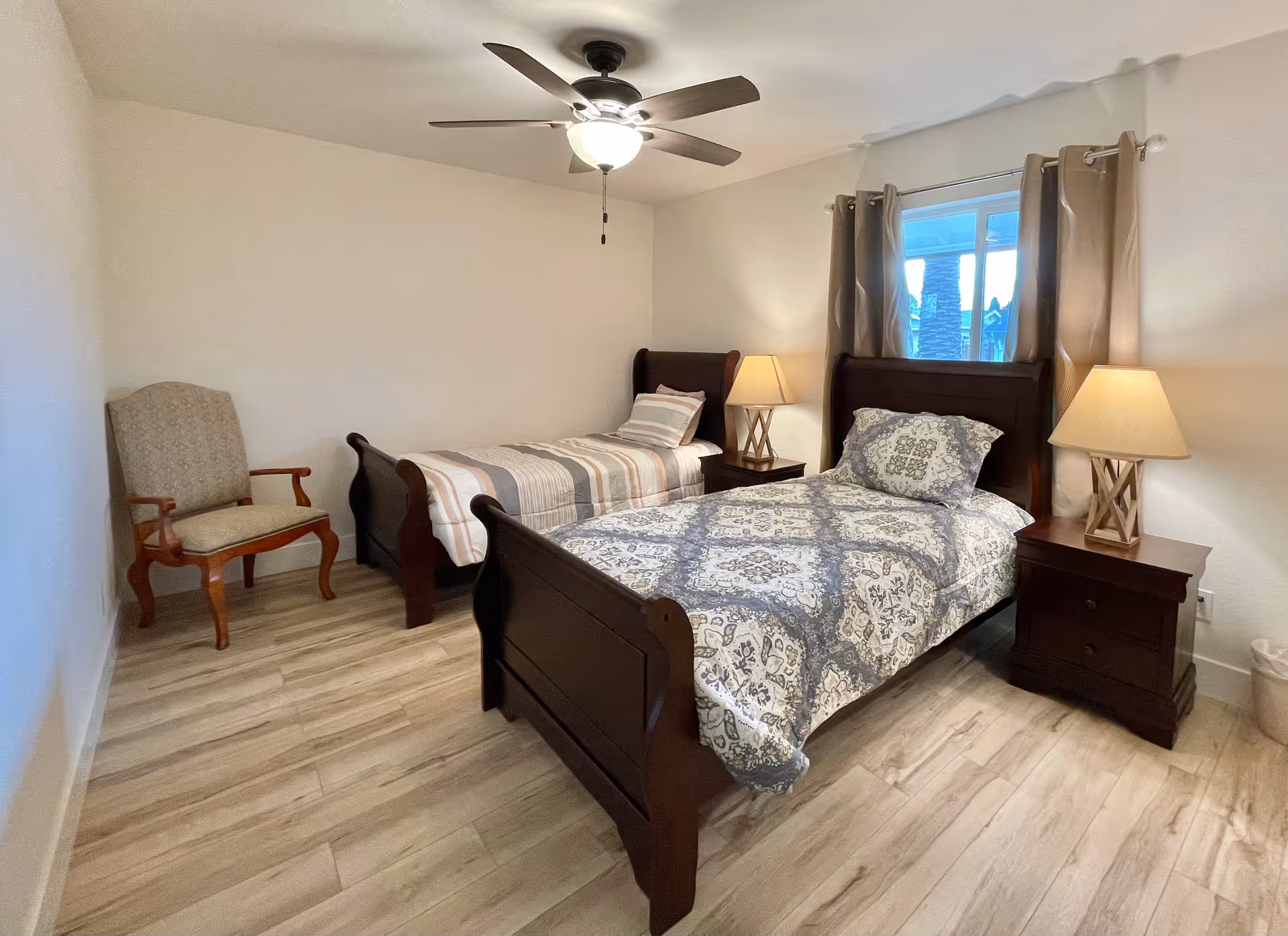 A bedroom with two single wooden beds, each with patterned bedding. There are two matching wooden nightstands with lamps on either side of the beds. A window with beige curtains is behind one of the beds. A ceiling fan with a light is mounted on the ceiling. There is also an upholstered wooden armchair in the corner of the room. The floor is light-colored wood.