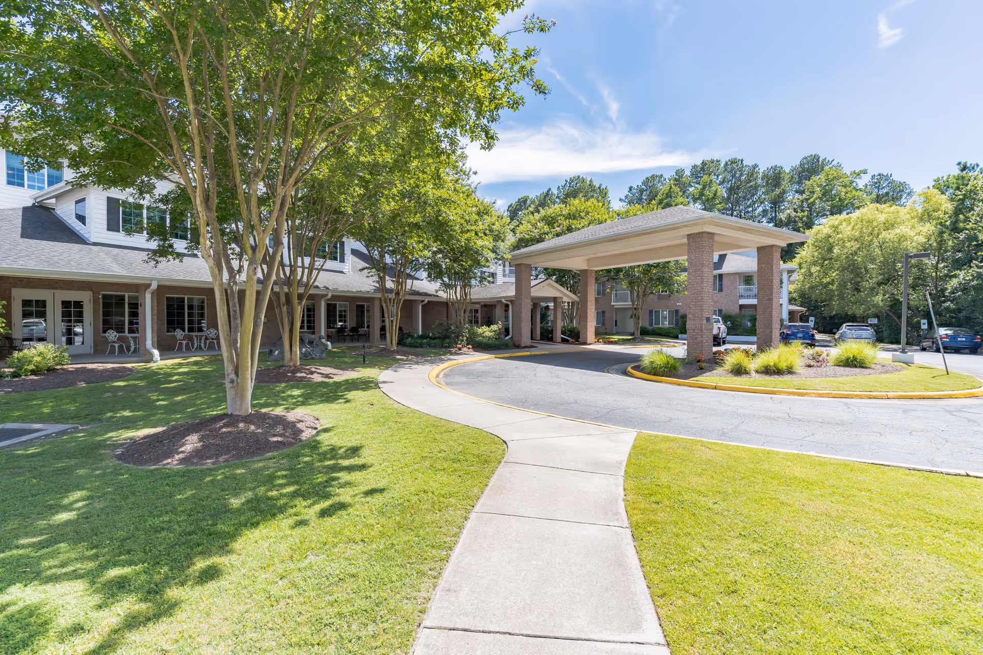 Exterior view of Lighthouse Pointe by Barclay House showing a covered entrance with brick pillars, a curved driveway, well-maintained green lawn, trees, and a parking area with several cars under a clear blue sky.