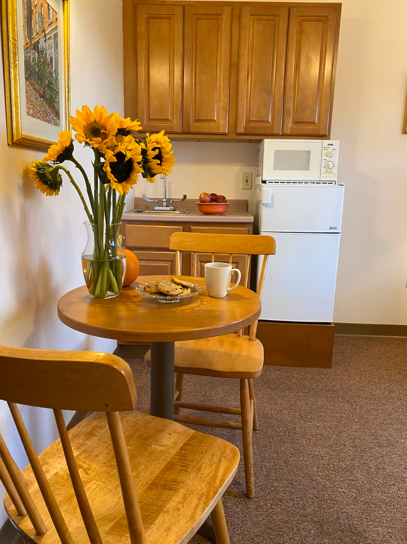 A small kitchenette and dining nook with a round wooden table and two chairs, sunflowers in a vase, a mug and cookies on the table, and cabinets with a mini fridge and microwave in the background.