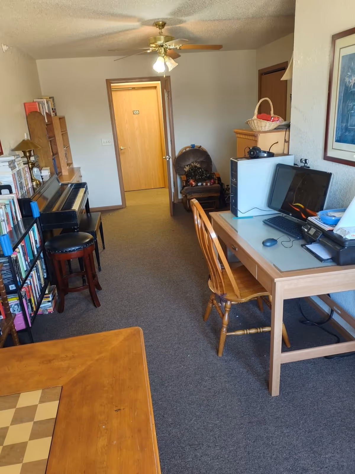 Interior room with a wooden desk holding a computer, keyboard, mouse, and printer on the right side. A wooden chair is placed in front of the desk. On the left side, there is a piano with a stool, a bookshelf filled with books, and a small table with a chessboard pattern. At the back, there is a rocking chair with some decorative items on it, and a closed wooden door labeled 103. The ceiling has a fan with lights.