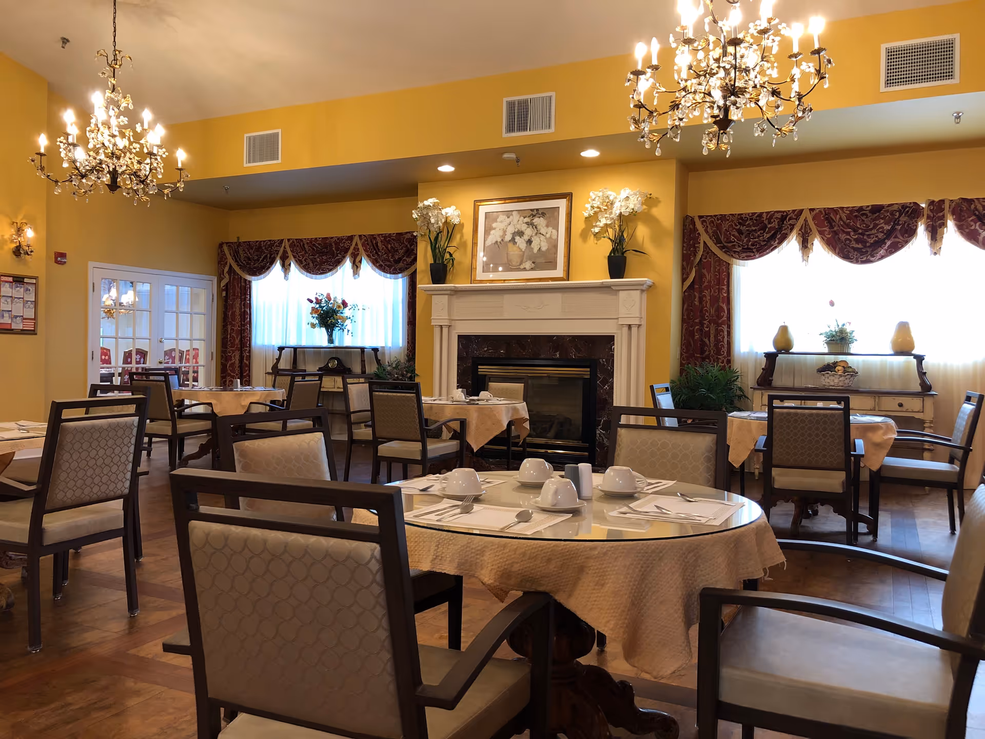 A dining room with multiple tables covered with beige tablecloths, each set with white cups, saucers, and silverware. The room has yellow walls, two ornate chandeliers, a white fireplace with floral artwork above it, and windows with red and gold curtains. There are decorative plants and flowers placed around the room.