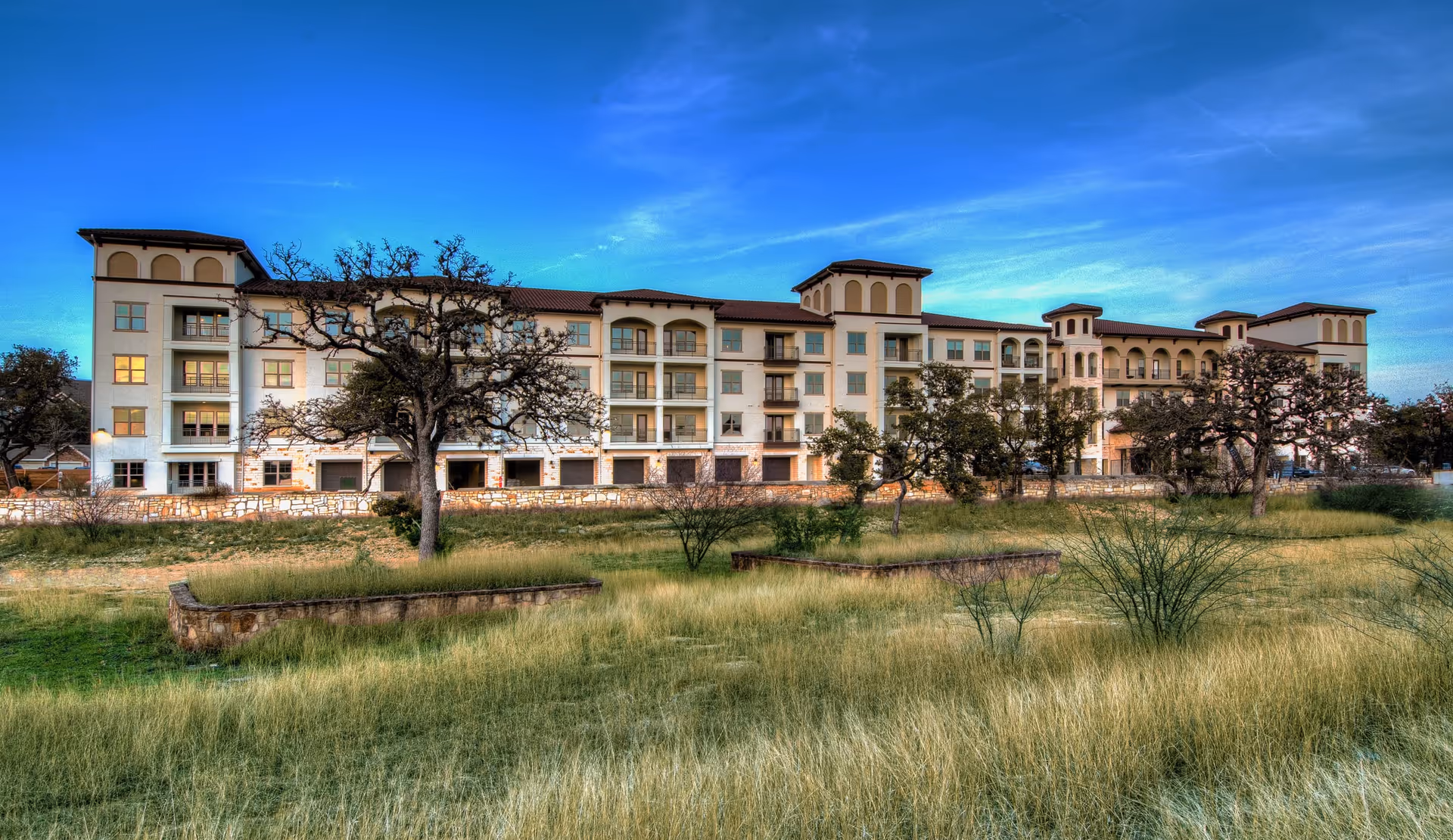 Multi-story Mediterranean-style senior living building with balconies overlooking grassy grounds and scattered trees under a blue sky.
