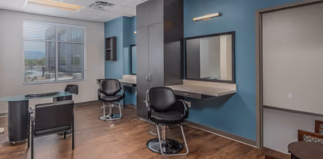 Interior view of a modern salon area with two black salon chairs in front of mirrors mounted on a blue wall. There is a small black table with two chairs near a window with blinds, and wooden flooring throughout the room.