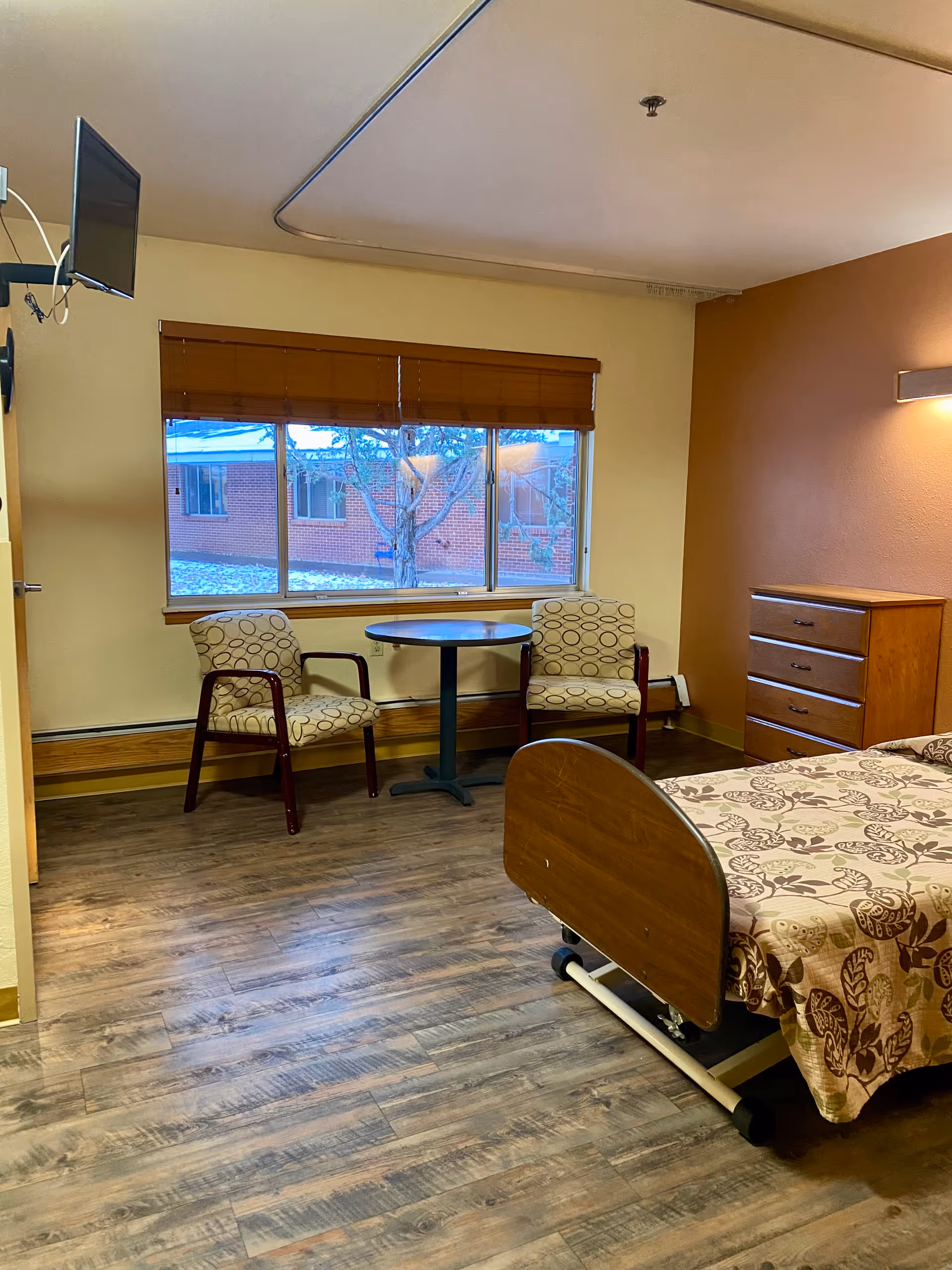 A furnished resident bedroom with a hospital-style bed, two chairs, a round table, and a dresser.