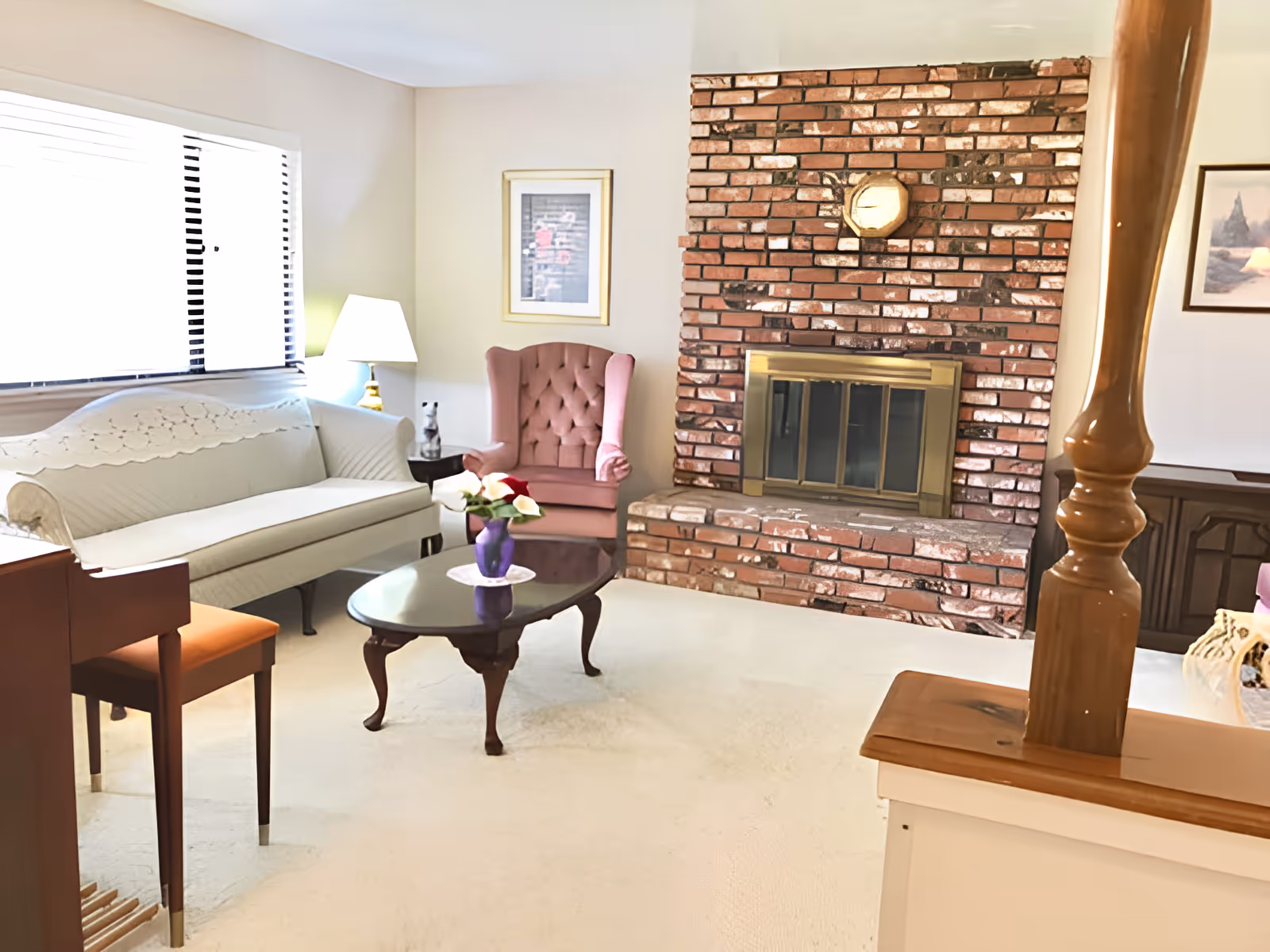 A bright living room with a brick fireplace, a white sofa, a pink armchair, and a wooden coffee table with a flower vase.
