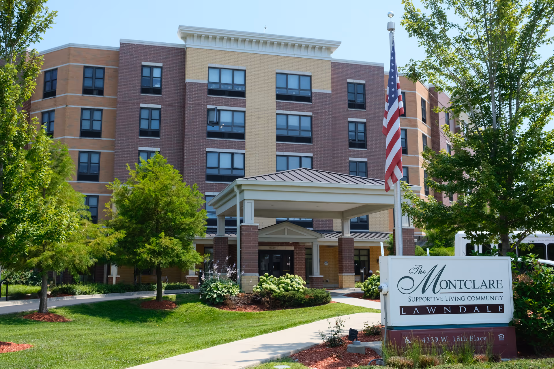 Front exterior of the Montclare supportive living community building with a covered entrance, landscaped lawn, American flag, and an entry sign.