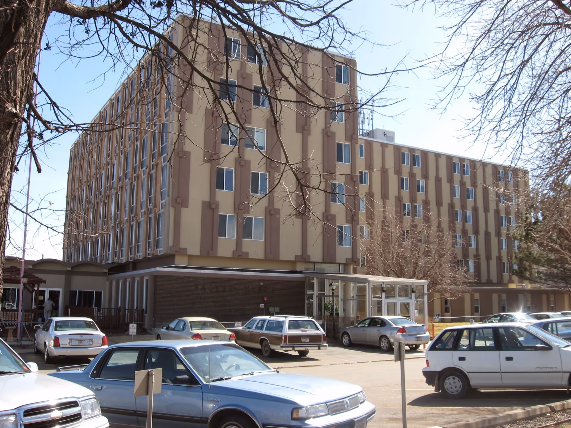 Exterior view of a multi-story retirement community building with several parked cars in front and leafless trees around the parking lot under a clear blue sky.