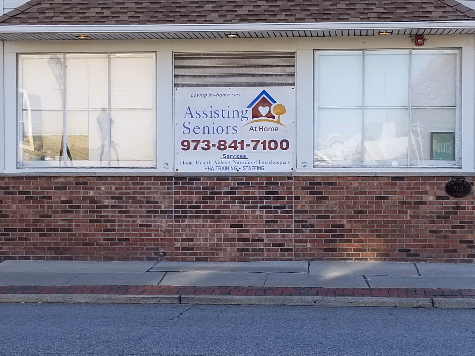 Brick storefront exterior with a banner sign reading Assisting Seniors At Home and a phone number.