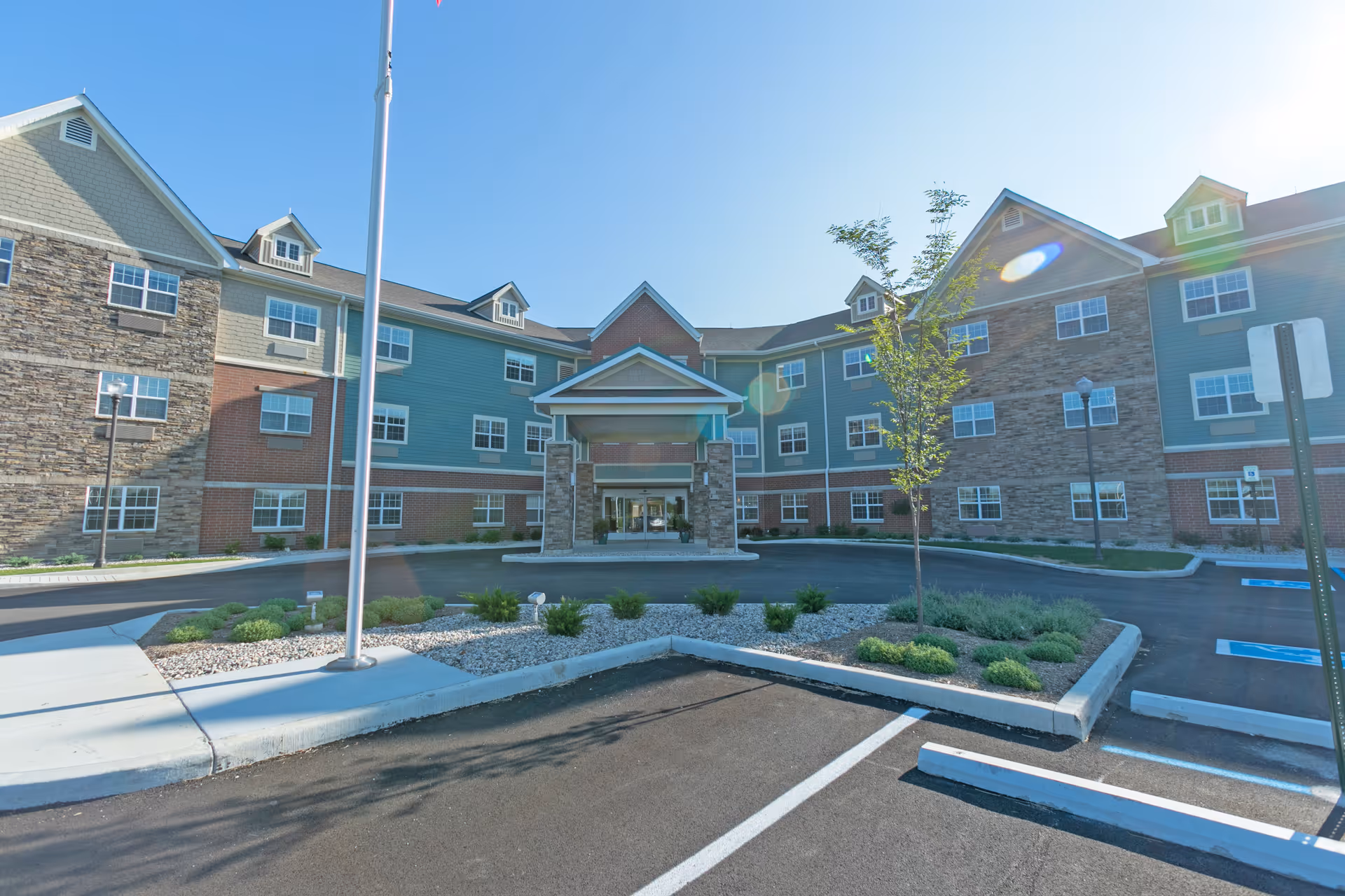 Exterior view of a senior living facility building with a covered entrance, multiple windows, and a mix of stone and siding on the facade. There is a parking lot with marked spaces and a landscaped area with small bushes and a young tree in front of the building under a clear blue sky.