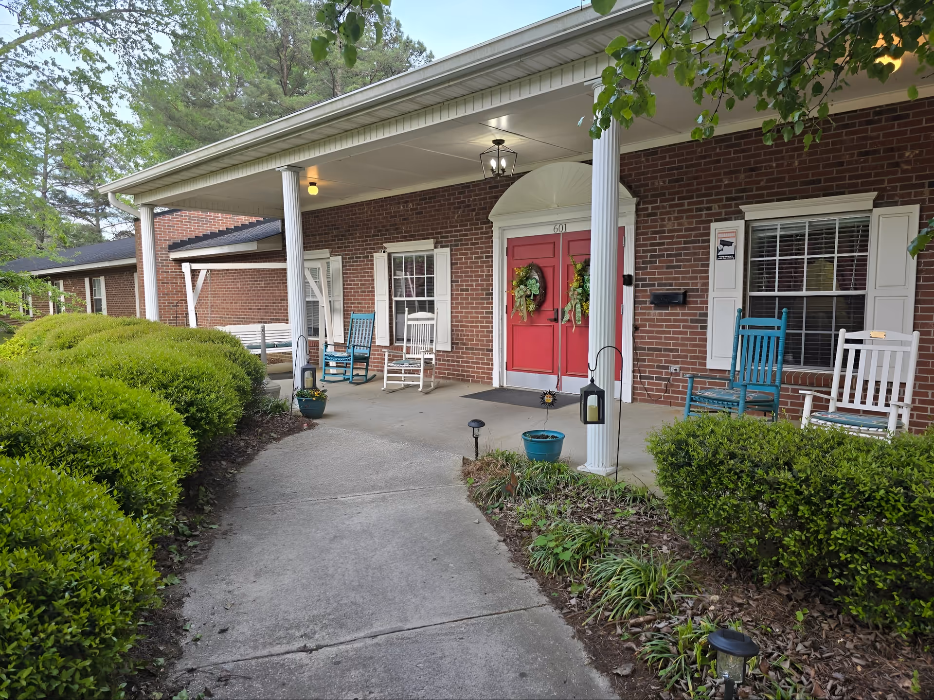 Brick building entrance with red double doors, white columns, and community rocking chairs on the porch.