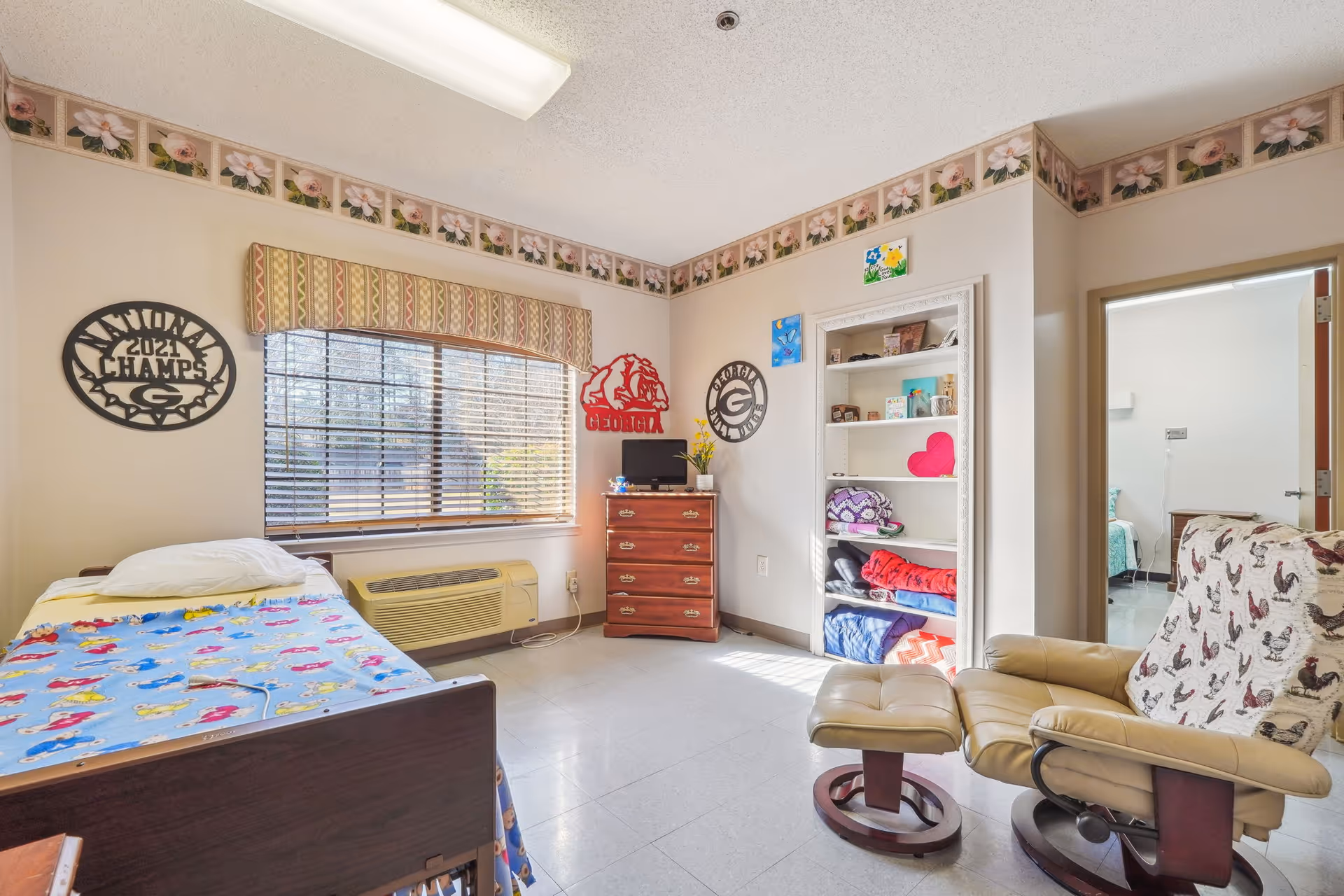 A cozy bedroom in a senior living facility with bed, chair, dresser, shelves, and window