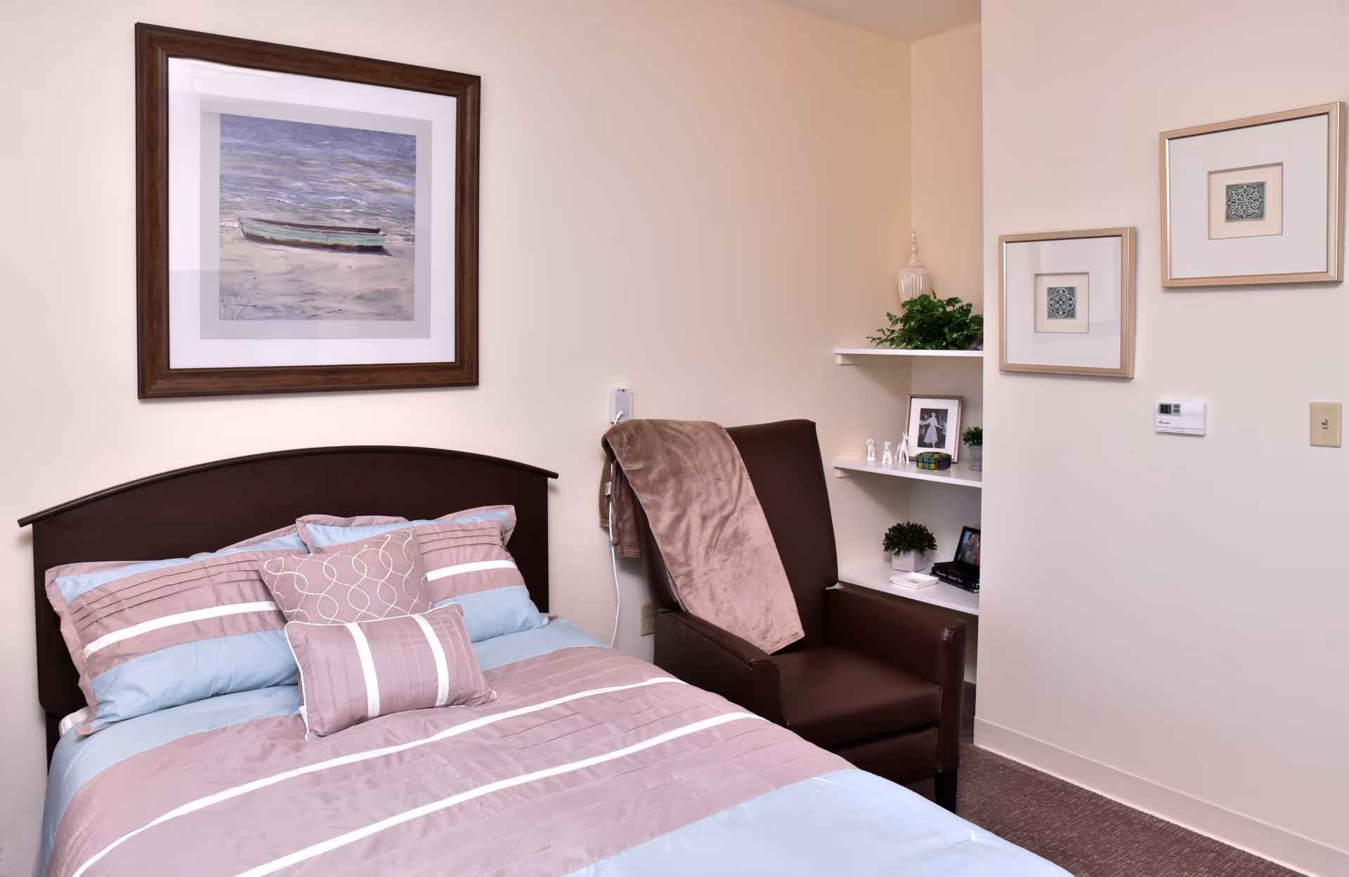 A small bedroom in a senior living facility with a bed, a brown chair, and decorative shelves on beige walls.