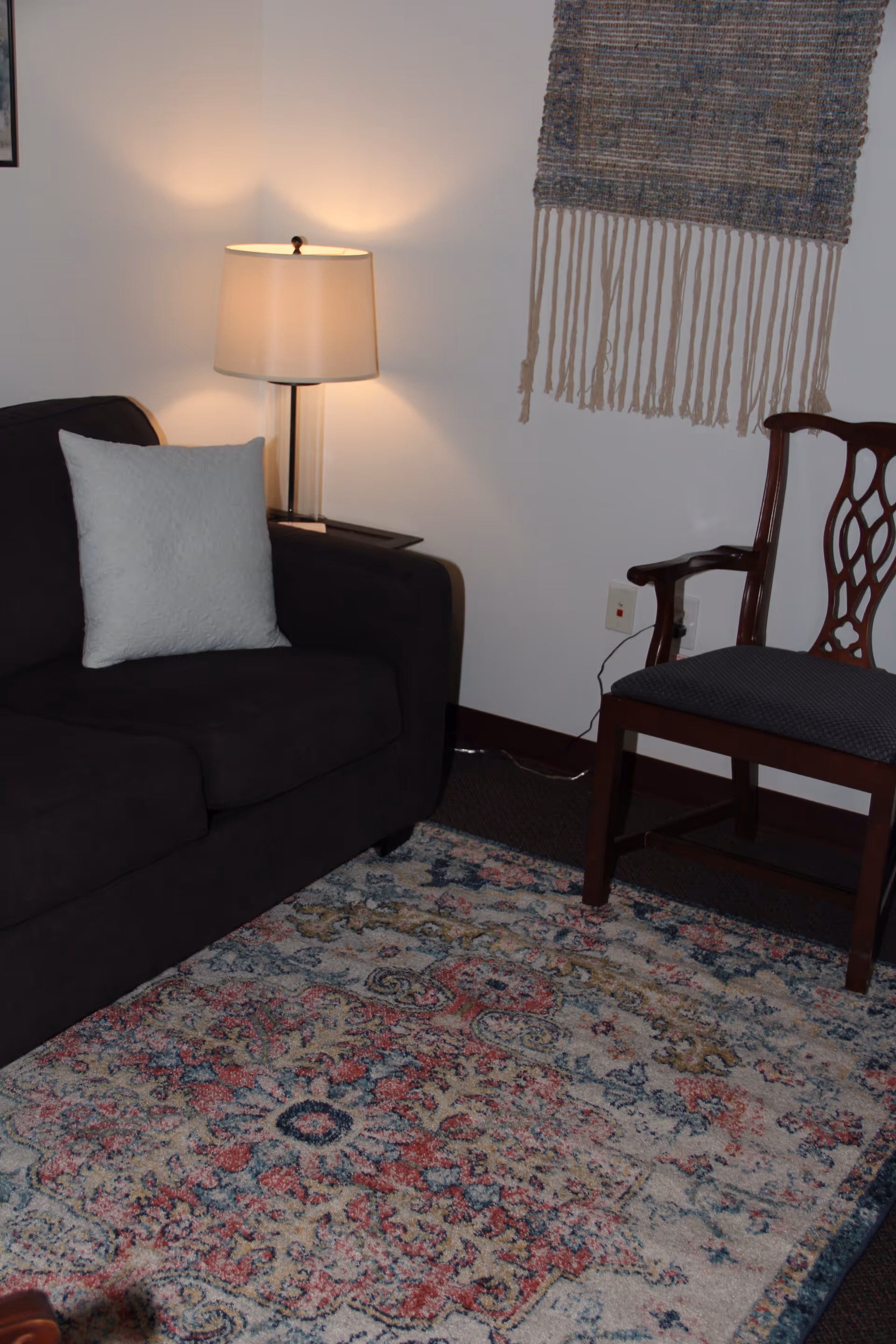 Cozy living room with dark sofa, patterned rug, lamp, and wooden chair.