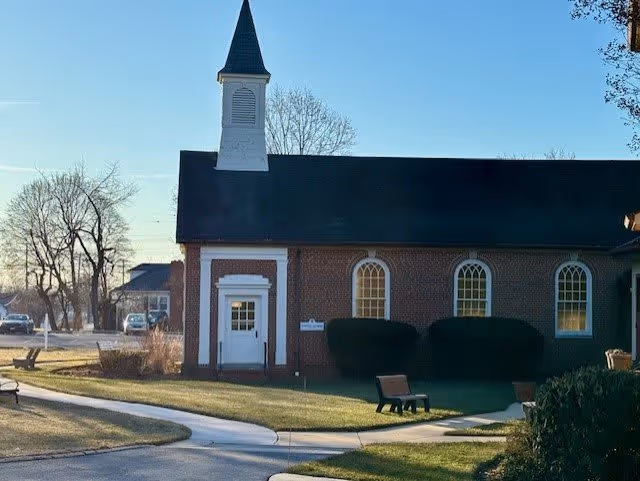 Brick chapel-like building with a small white steeple, arched windows, and a walkway across a lawn in front.