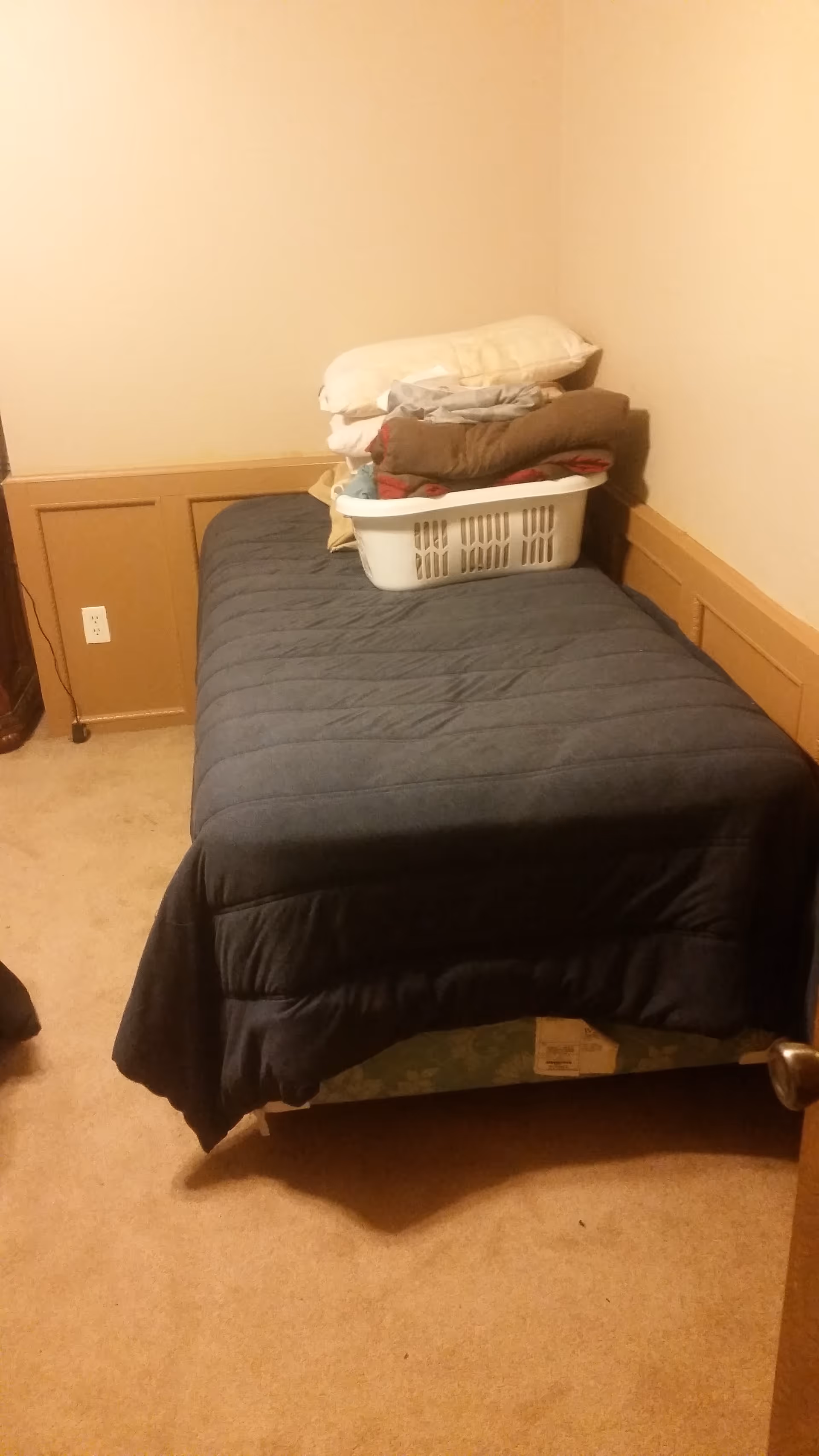 A small bedroom with a single bed, a dark quilt, and a laundry basket on it.