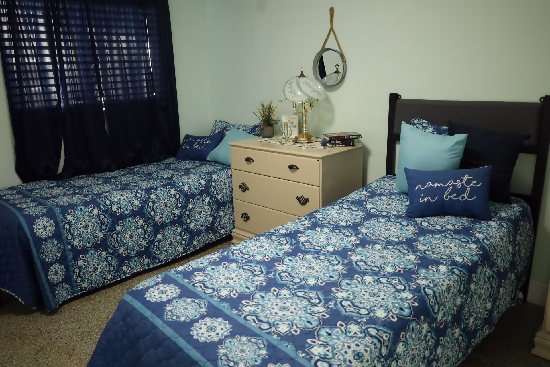 Two twin beds with blue patterned quilts, a nightstand, and dark curtains.