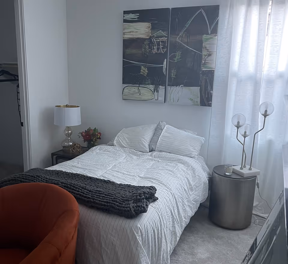 A neatly made bedroom with white bedding, abstract wall art, and a metal bedside table.