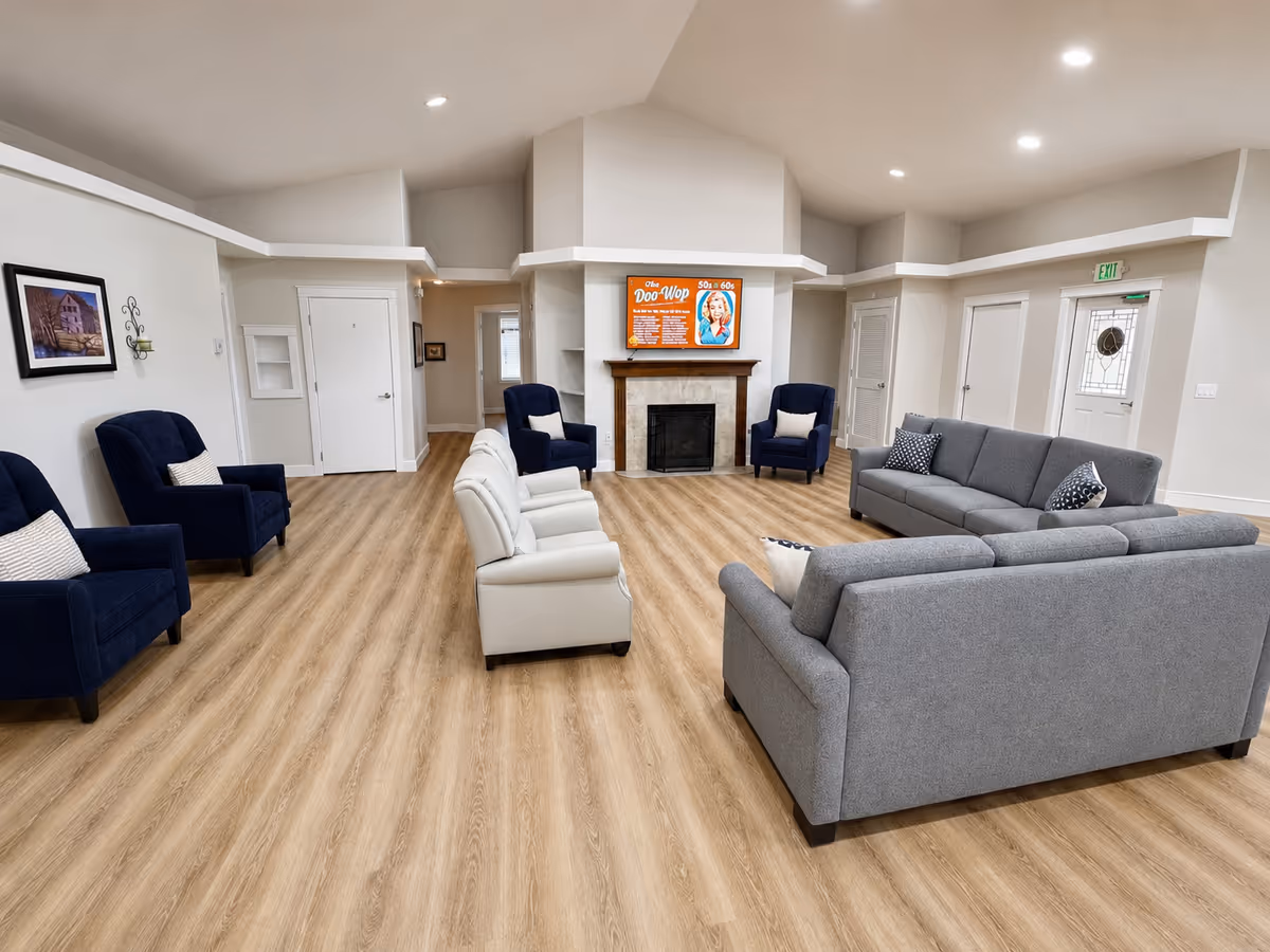 Spacious senior living lounge with gray sofas, navy chairs, and a central fireplace.