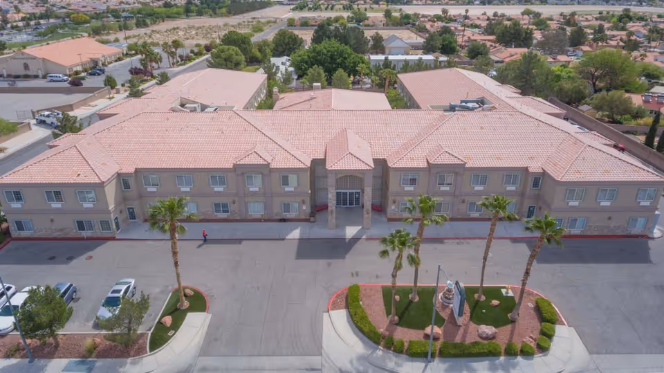 Aerial view of Desert View Senior Living facility showing a large, two-story building with a tiled roof, surrounded by palm trees and landscaped areas. The building has multiple windows and a central entrance with columns. There are parked cars and paved driveways in front of the building.