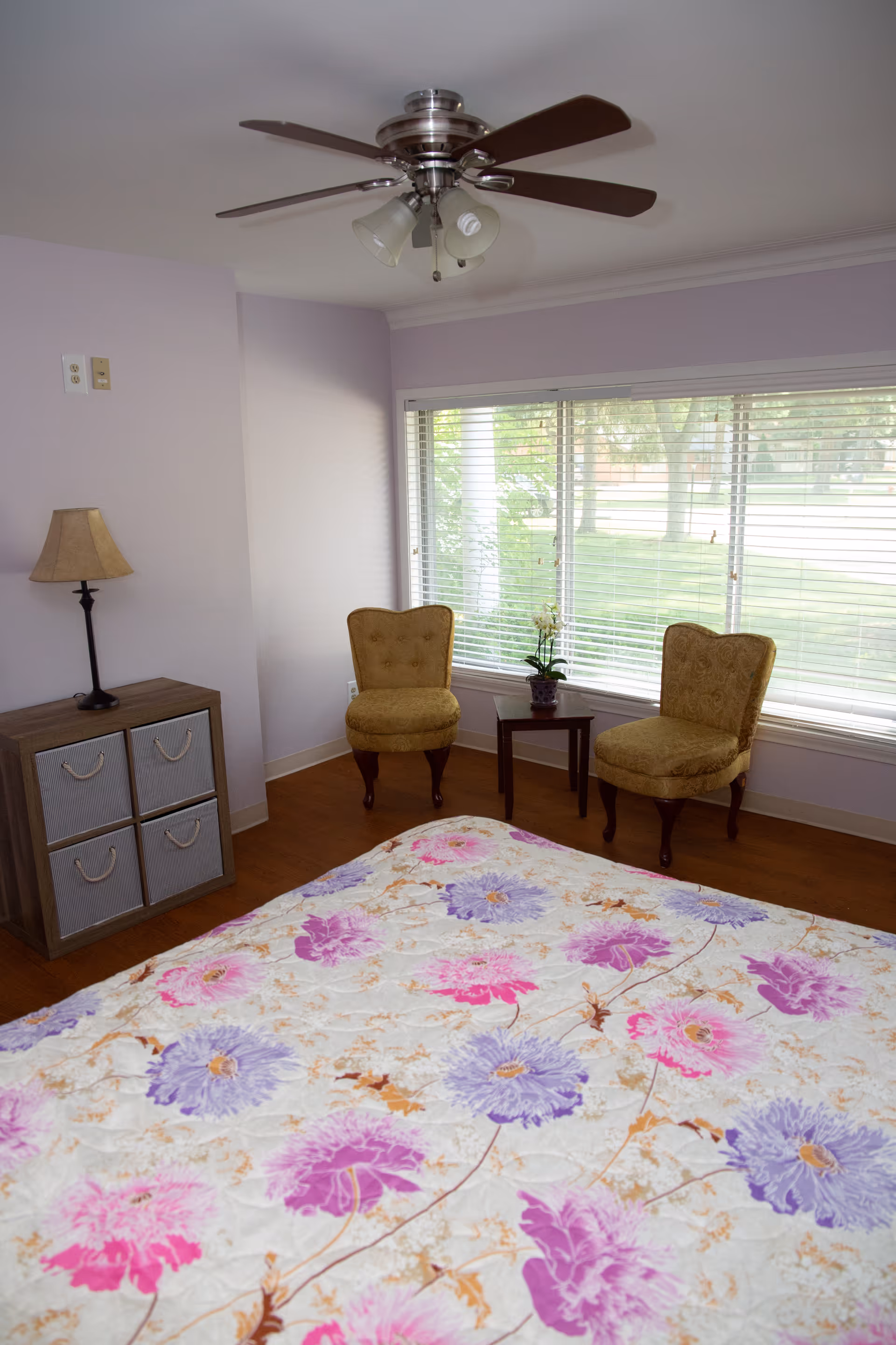 A cozy bedroom featuring a floral bedspread, two chairs by a window, and a ceiling fan.