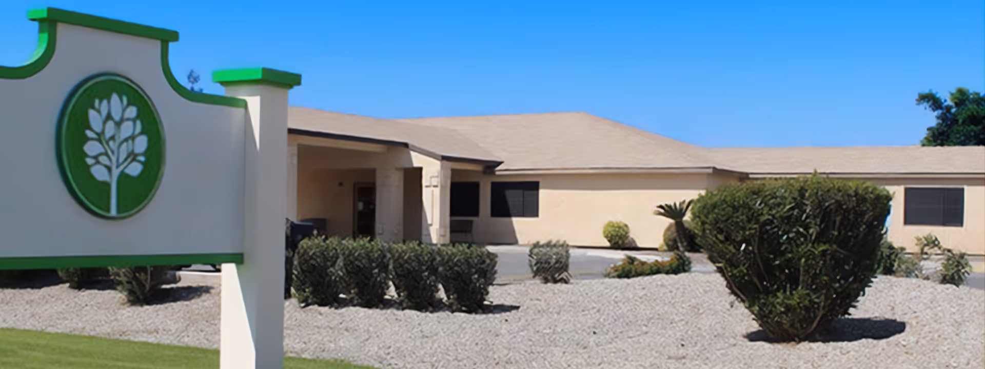 Exterior view of a single-story beige building with a tan roof under a clear blue sky. In the foreground, there is a landscaped area with gravel and trimmed bushes. A white and green signpost with a tree emblem is visible on the left side of the image.