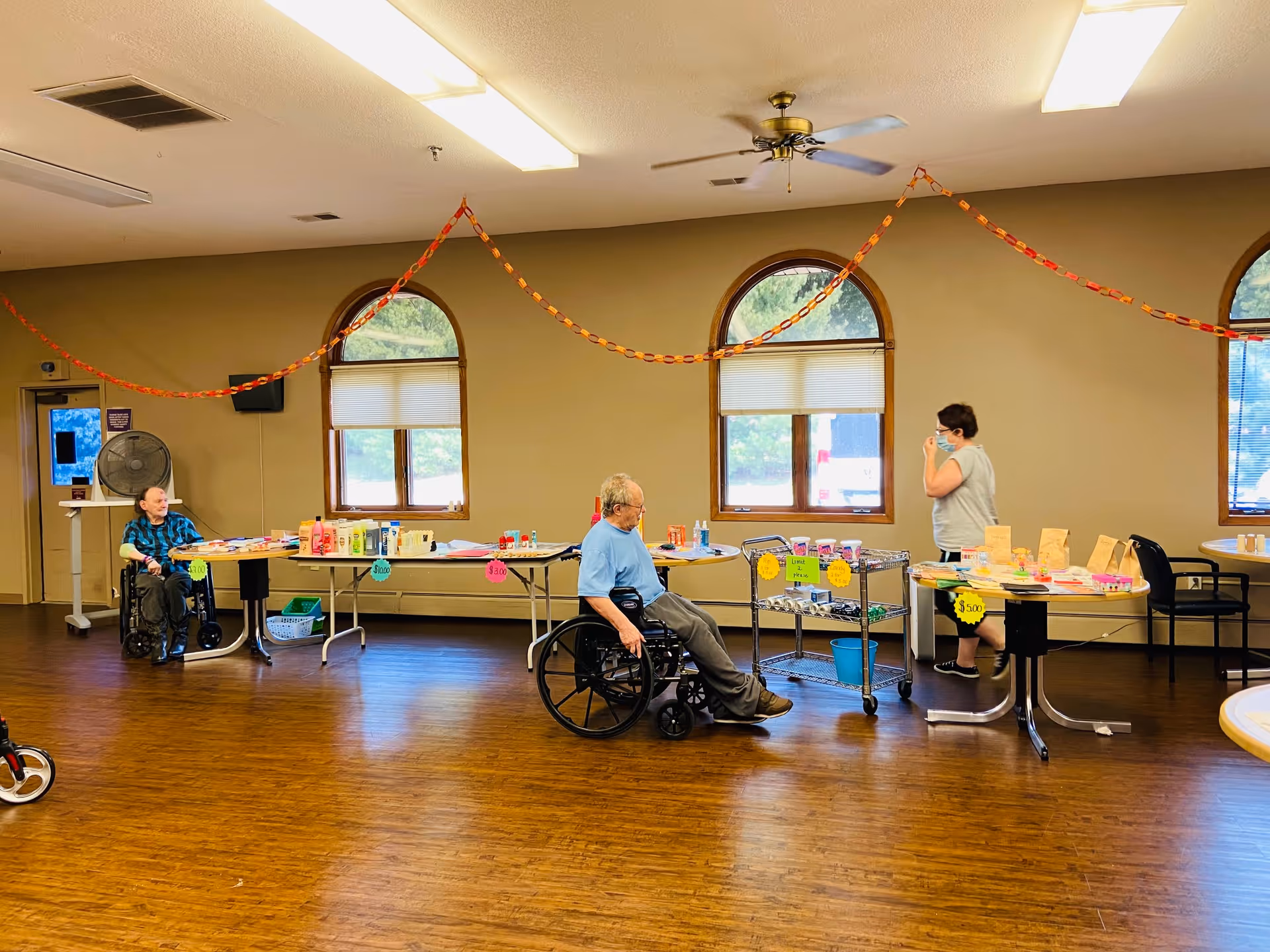 A spacious room with wooden flooring and beige walls featuring three arched windows. Two elderly individuals in wheelchairs are positioned near tables displaying various items with price tags. A woman wearing a mask stands near one of the tables. Orange and red paper chains are hung across the room as decorations.