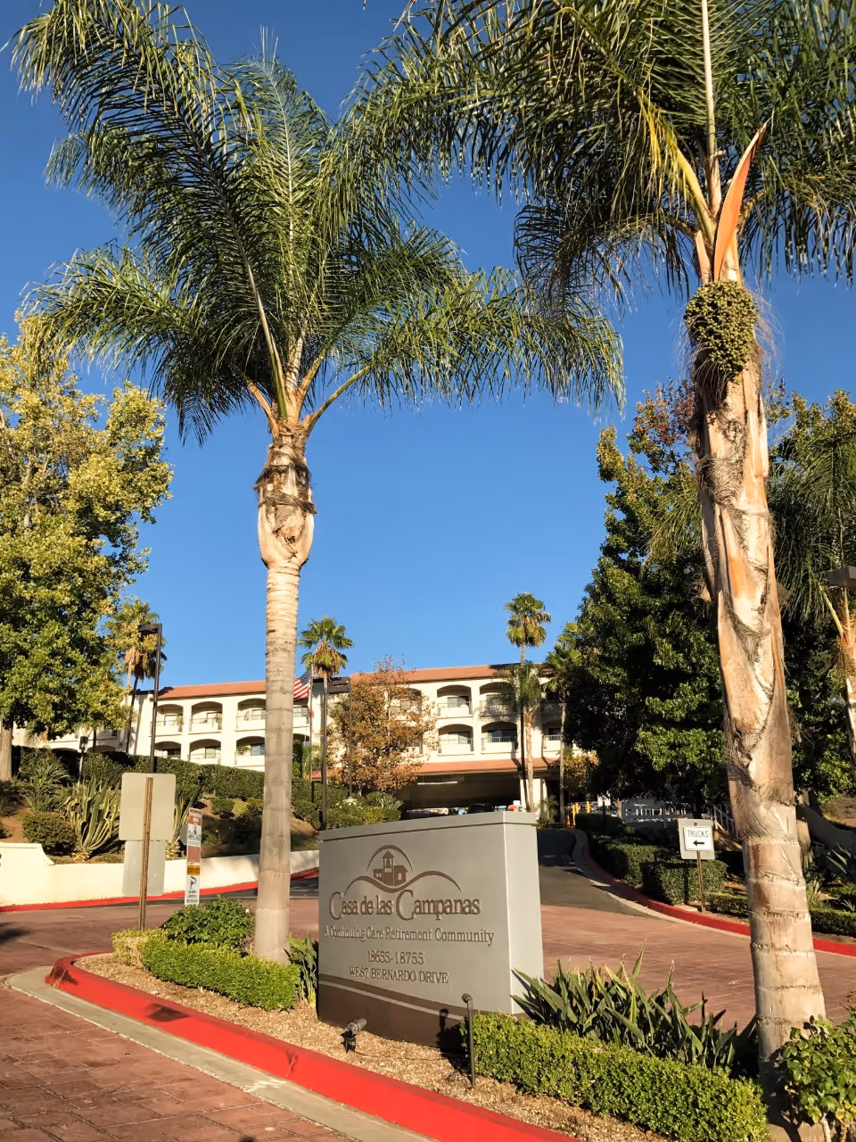 Entrance to Casa de las Campanas, a continuing care retirement community, with a large sign displaying the facility name and address. The driveway is lined with palm trees and other greenery, and the multi-story building is visible in the background under a clear blue sky.