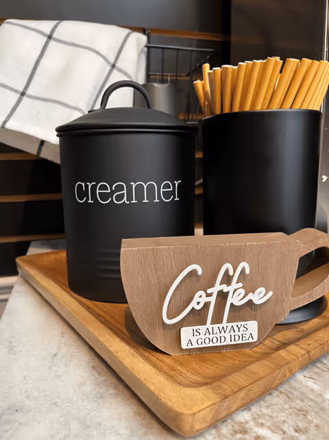 A wooden tray on a countertop holding a black container labeled 'creamer', a black cup filled with brown paper straws, and a wooden coffee cup-shaped sign that reads 'Coffee is always a good idea'. A white cloth with a black grid pattern is hanging in the background.