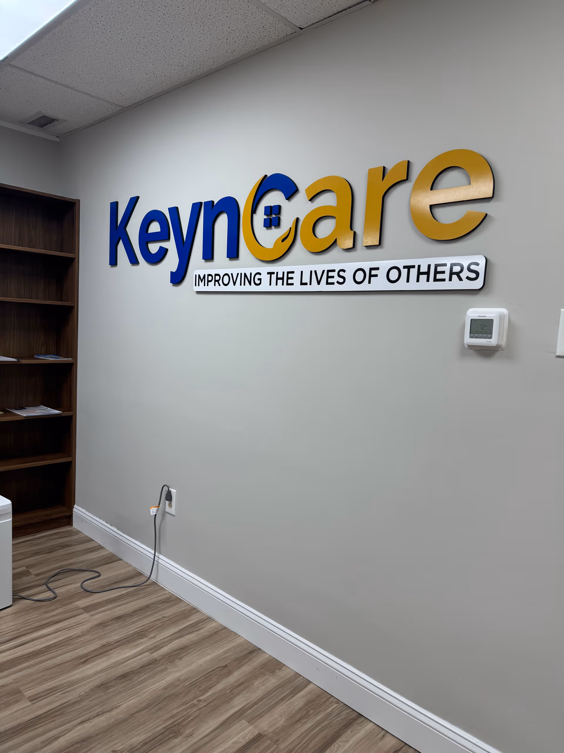 Interior wall featuring KeynCare logo and slogan, with a bookshelf and wood floor.