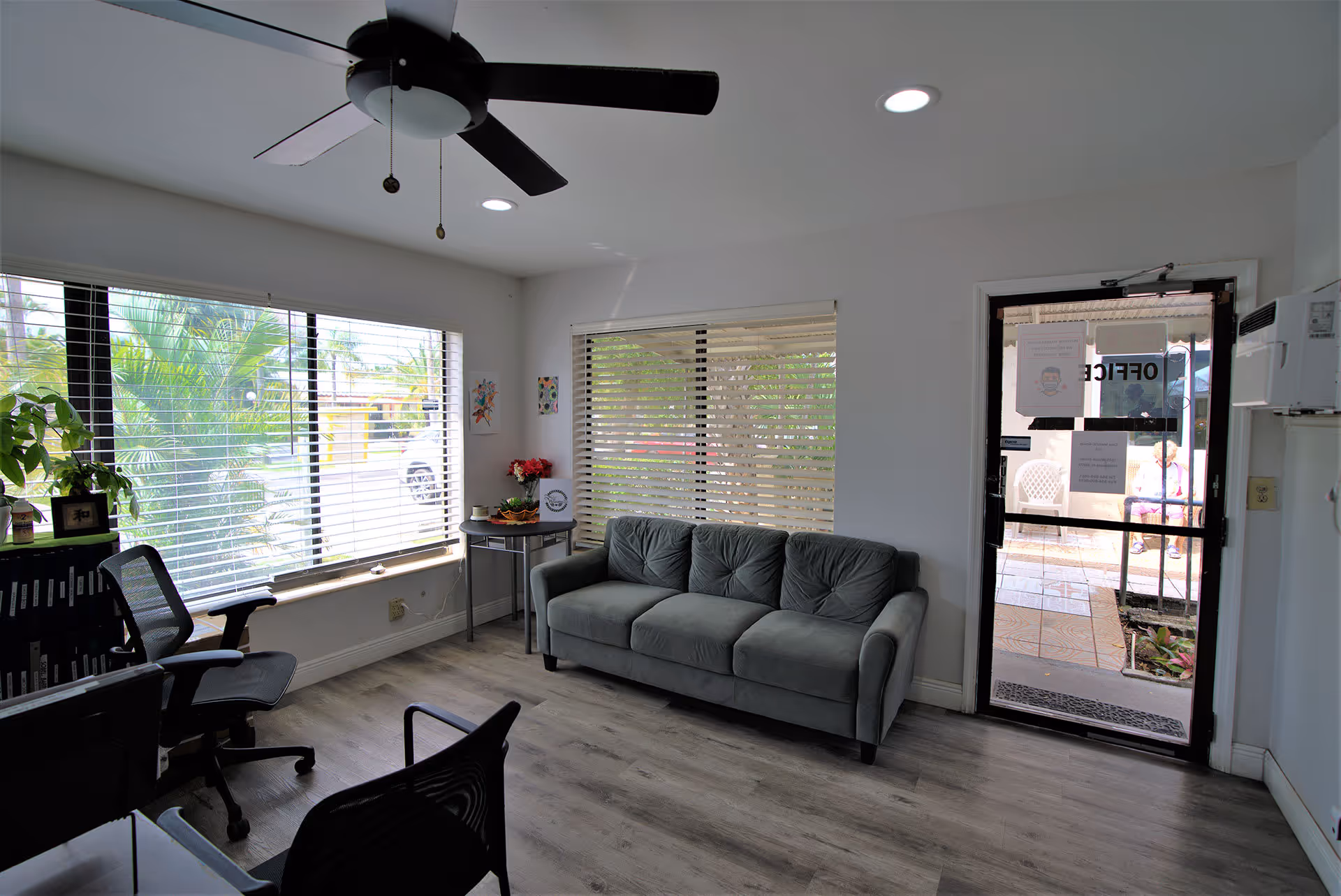 A small office or waiting area with a gray couch, two black office chairs, a ceiling fan, and large windows with blinds. Outside the glass door labeled 'OFFICE,' a person is sitting on a white chair on a tiled patio.