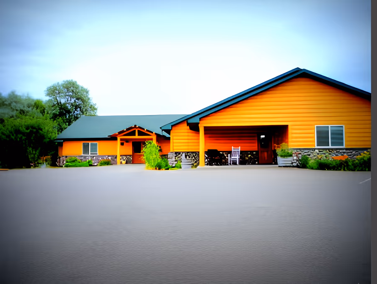 Single-story orange building with a covered entrance, rocking chairs, landscaping, and a large empty parking area in front.