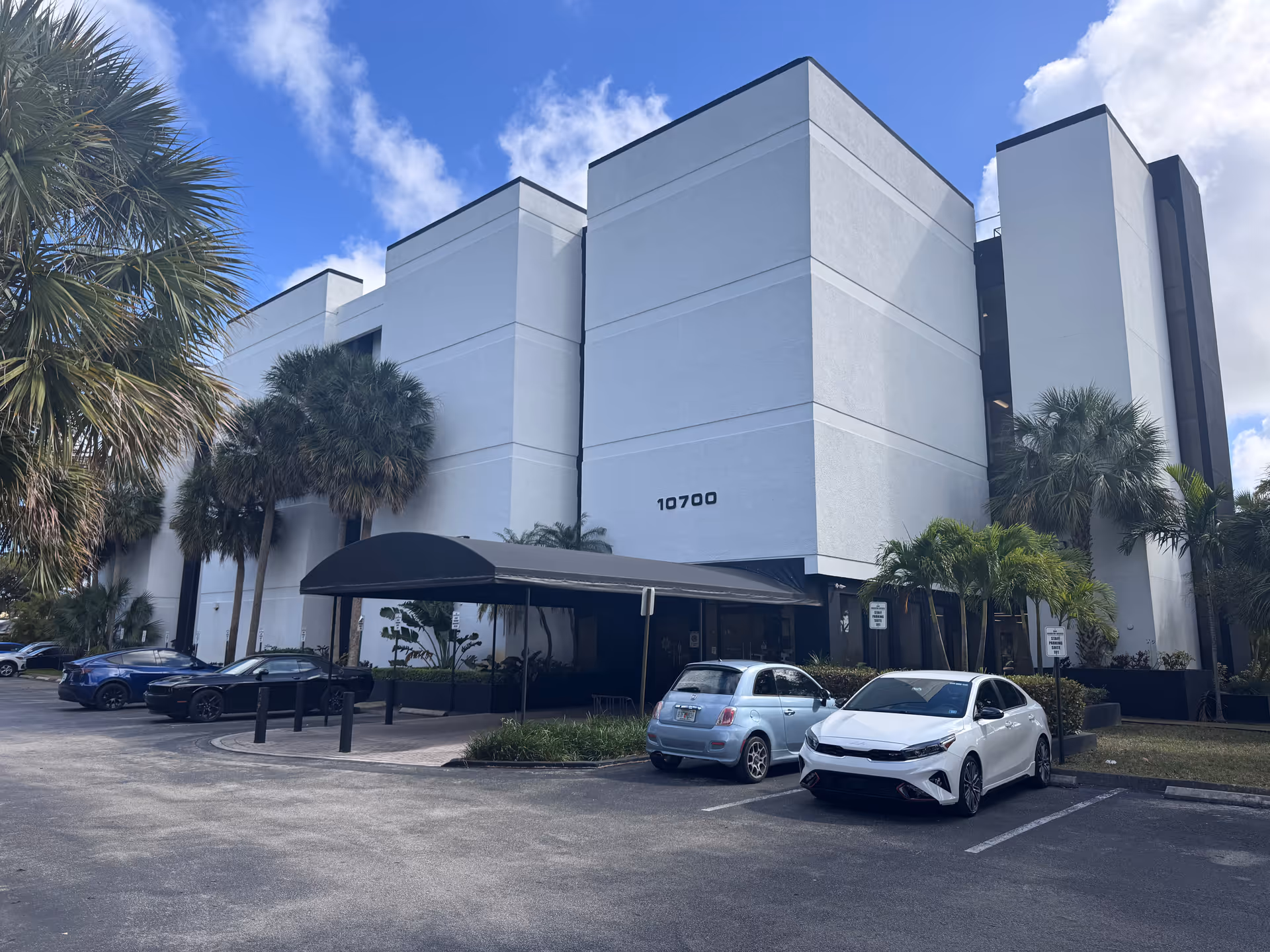 A modern white senior living facility exterior with a covered entrance and palm trees.