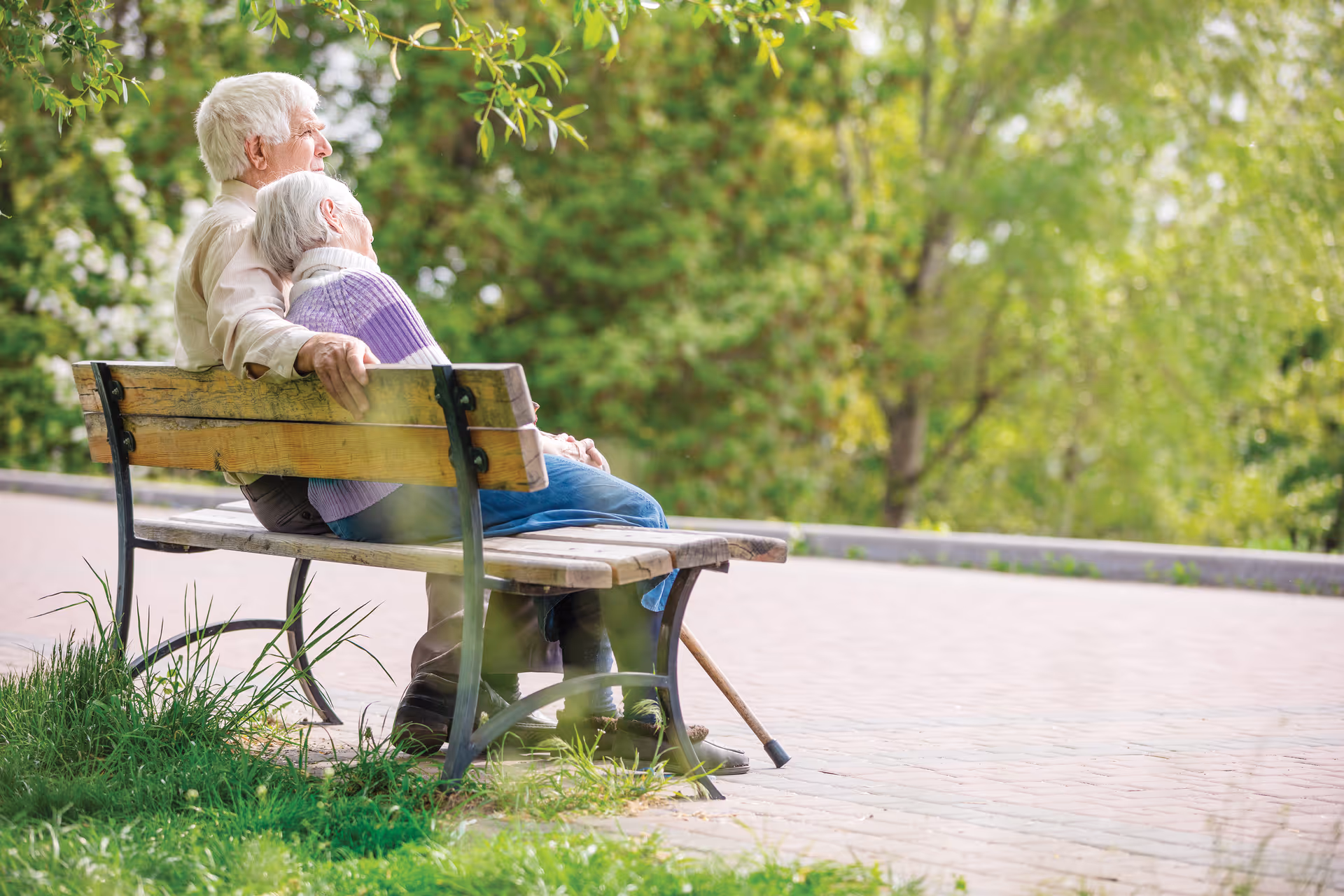 Two elderly residents sit on a wooden bench in a sunlit park.